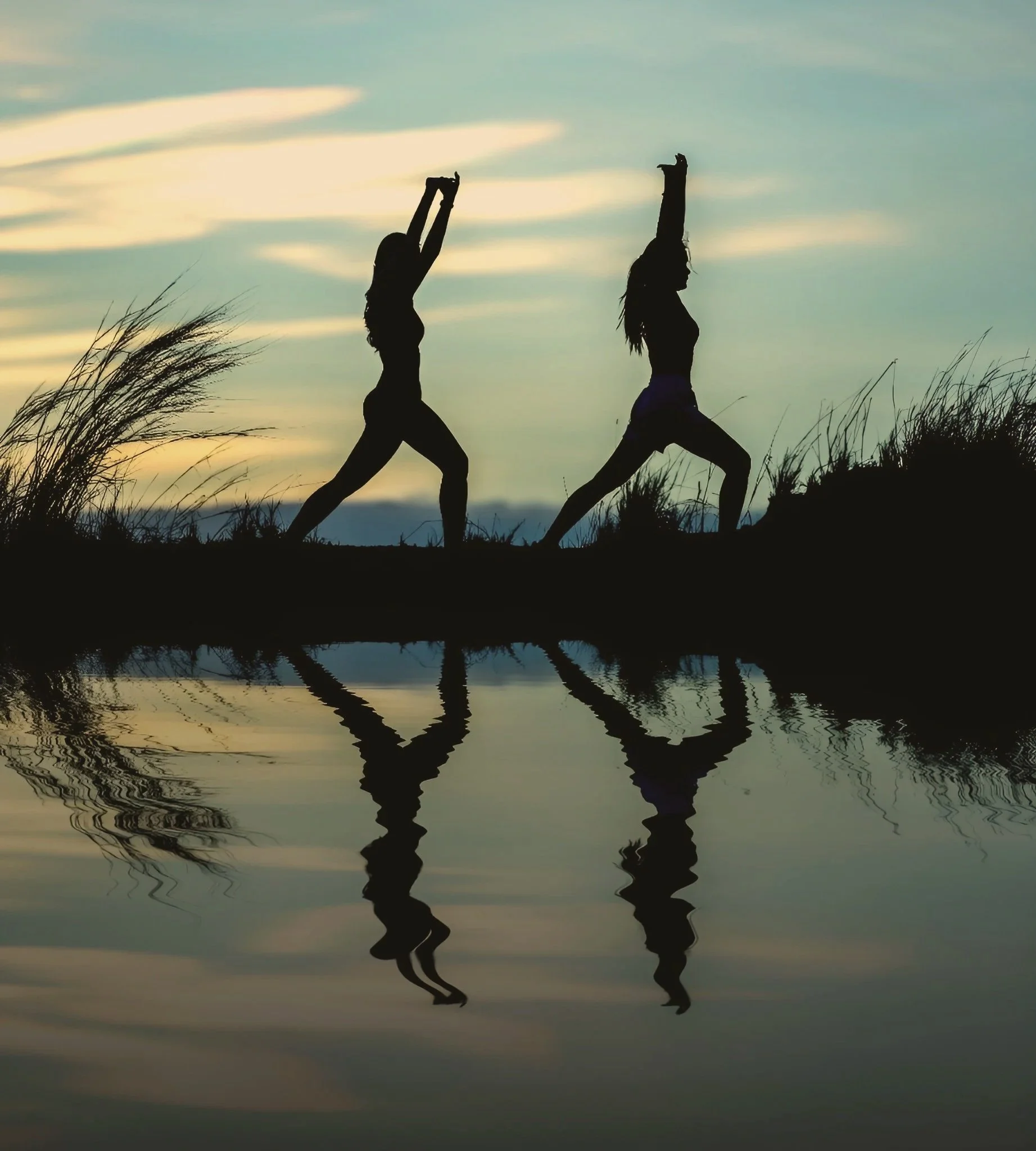 Silhouettes of two women practicing yoga outdoors during sunset, with their reflections visible in a body of water.
