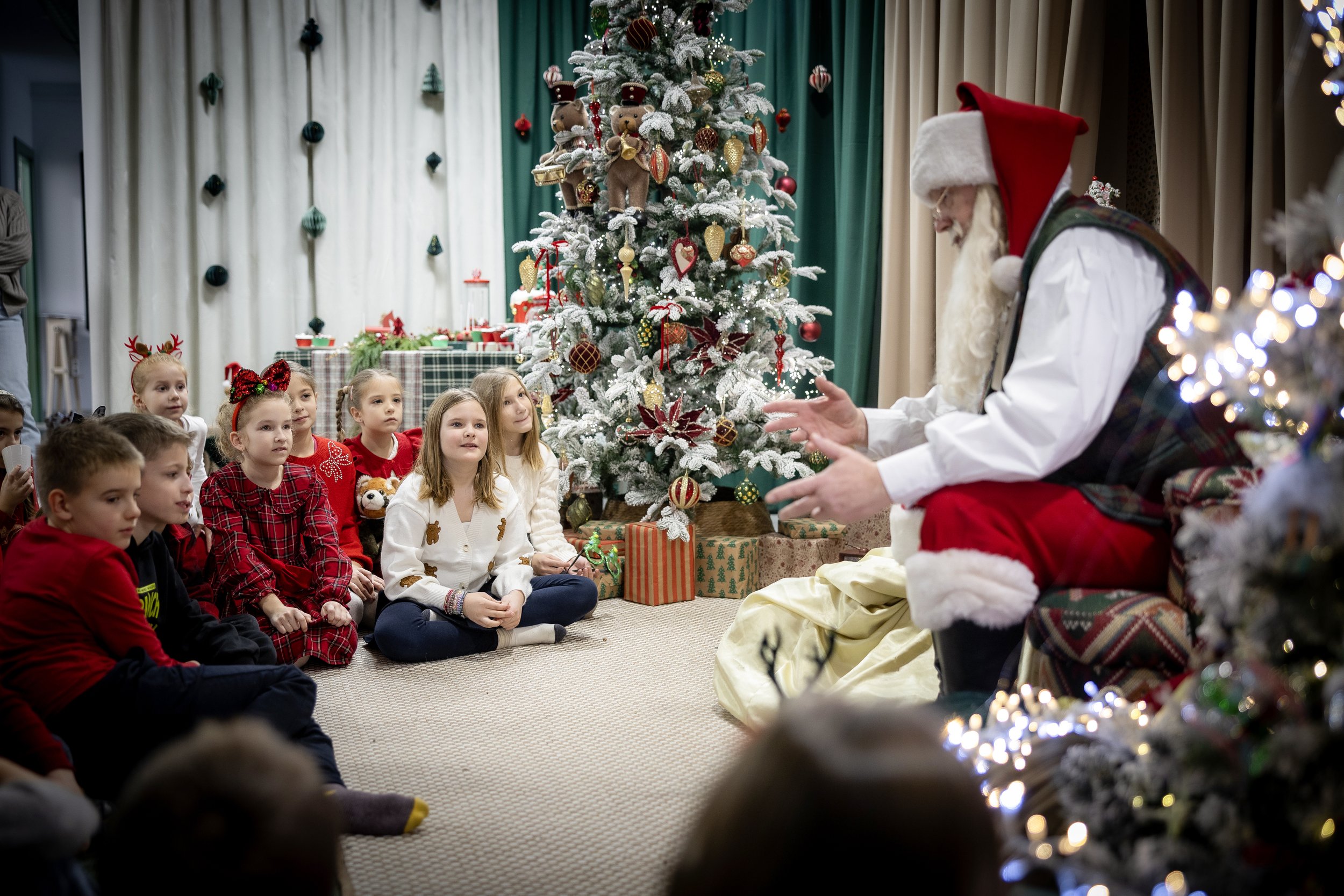 Children sitting on the floor watching a man dressed as Santa Claus during a Christmas celebration with a decorated Christmas tree in the background.