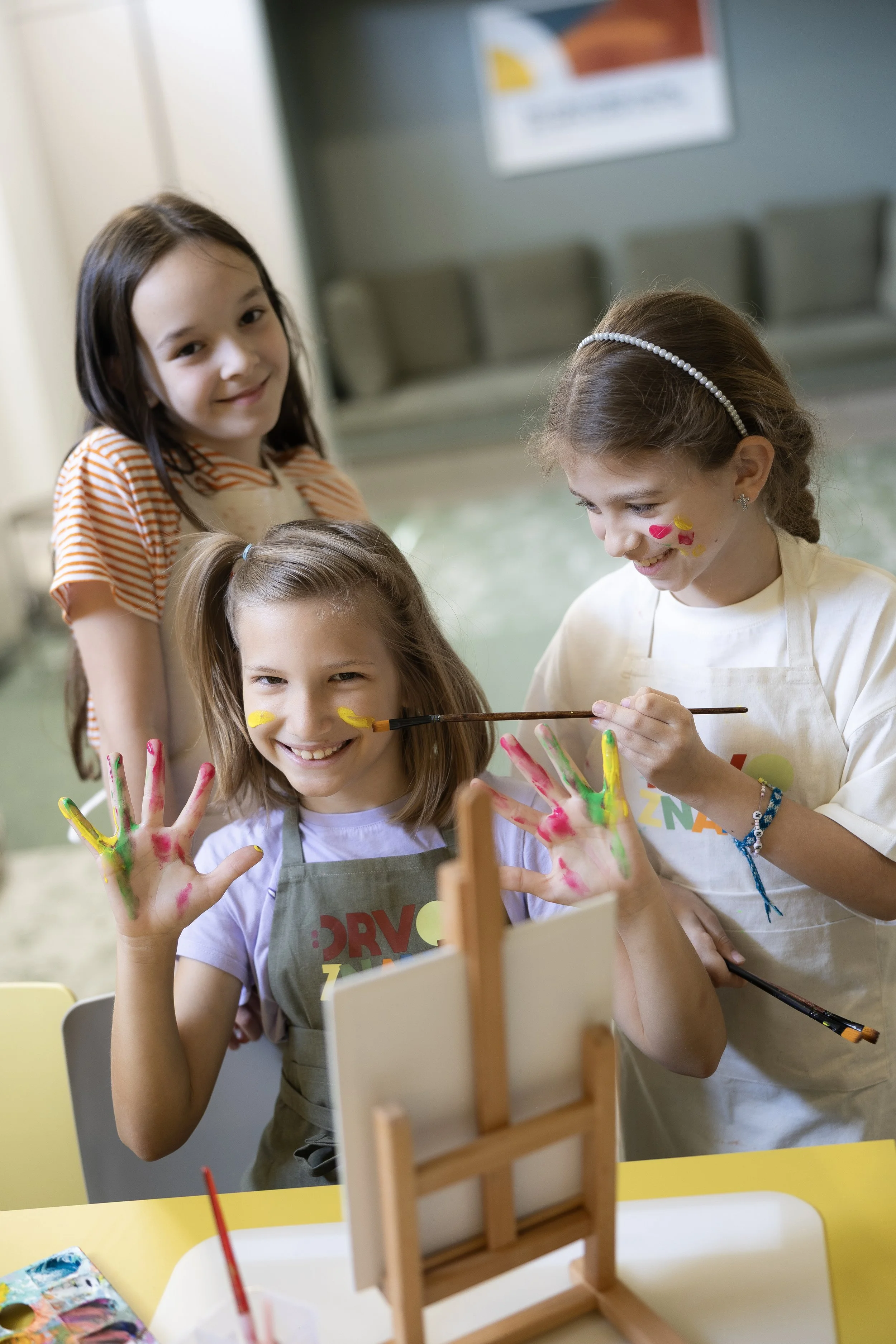 Children at an art activity with painted hands, smiling and with face paint, in an indoor setting.