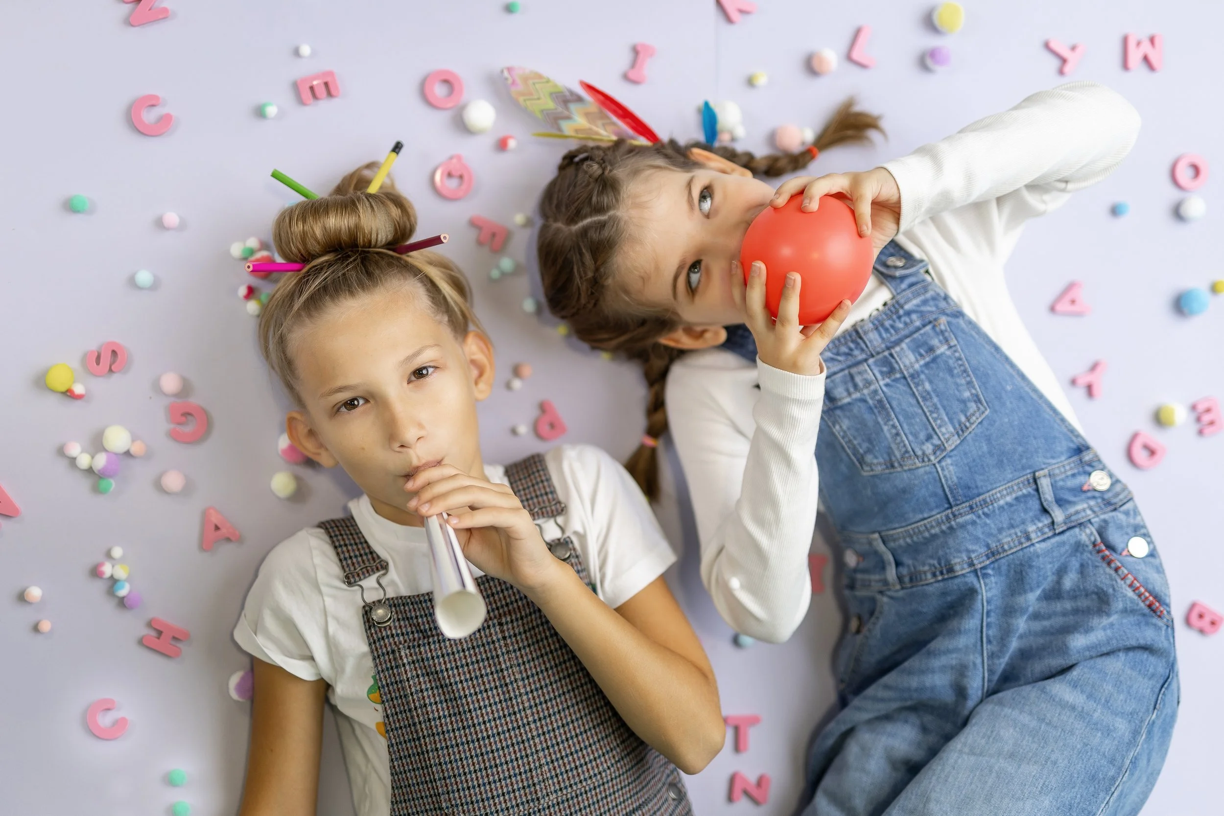 Two young girls lying on the floor surrounded by colorful alphabet letters, pom-pom decorations, and confetti, one girl holding a red balloon and the other with a party horn.