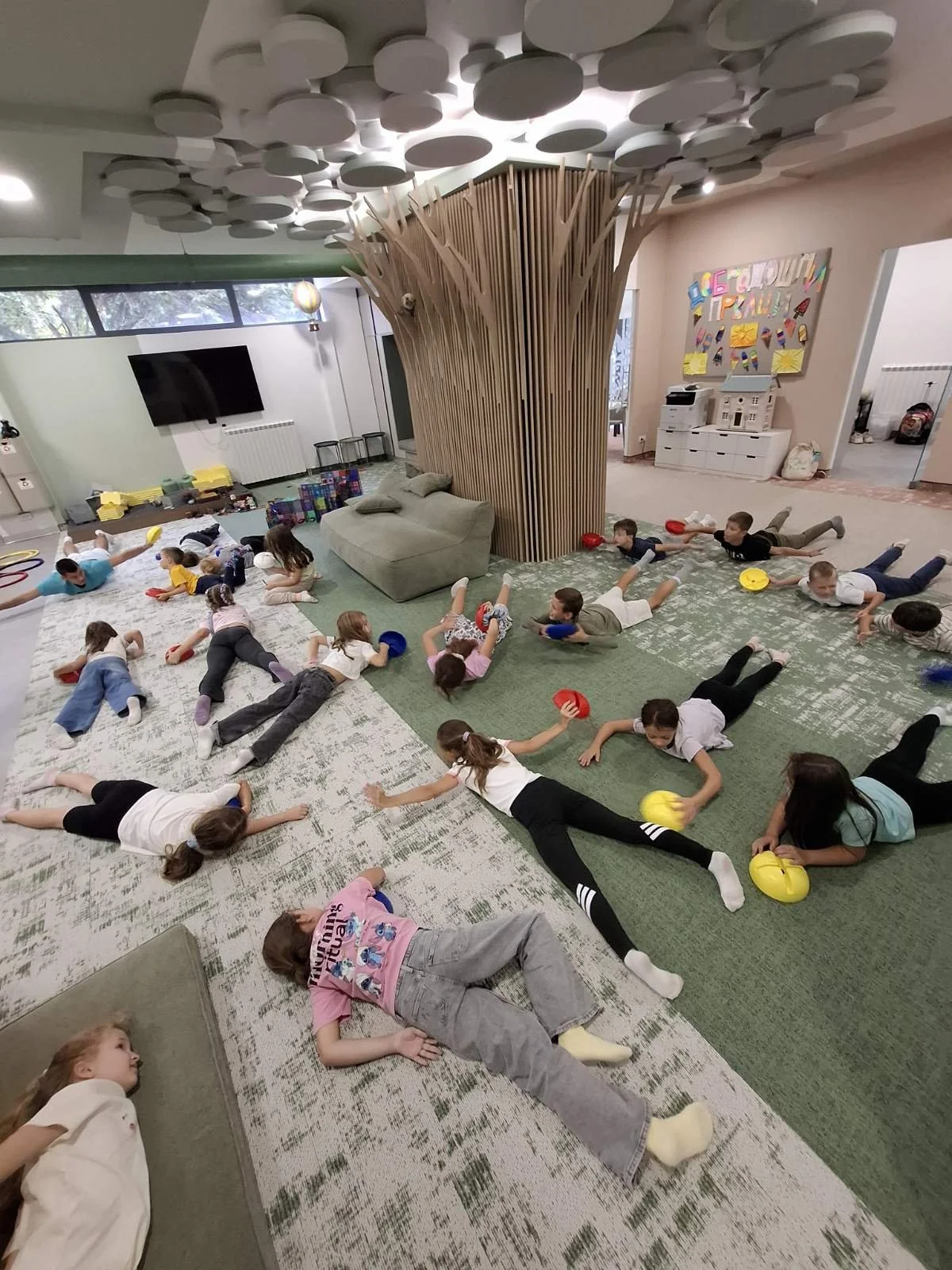 Children lying on the floor in a playroom, some holding colorful balls, with a wooden tree-like structure and a large gray couch in the background.