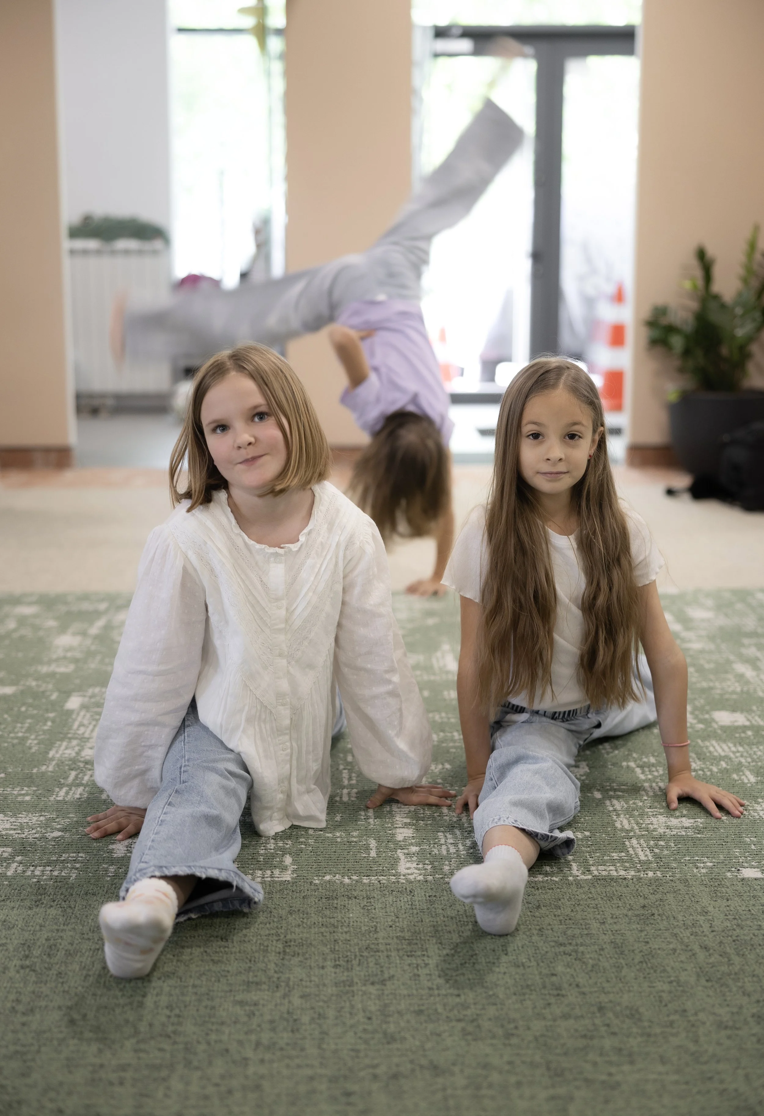 Two young girls sitting on a green patterned carpet in a room with beige walls, with a third girl in the background doing a handstand against a glass door.