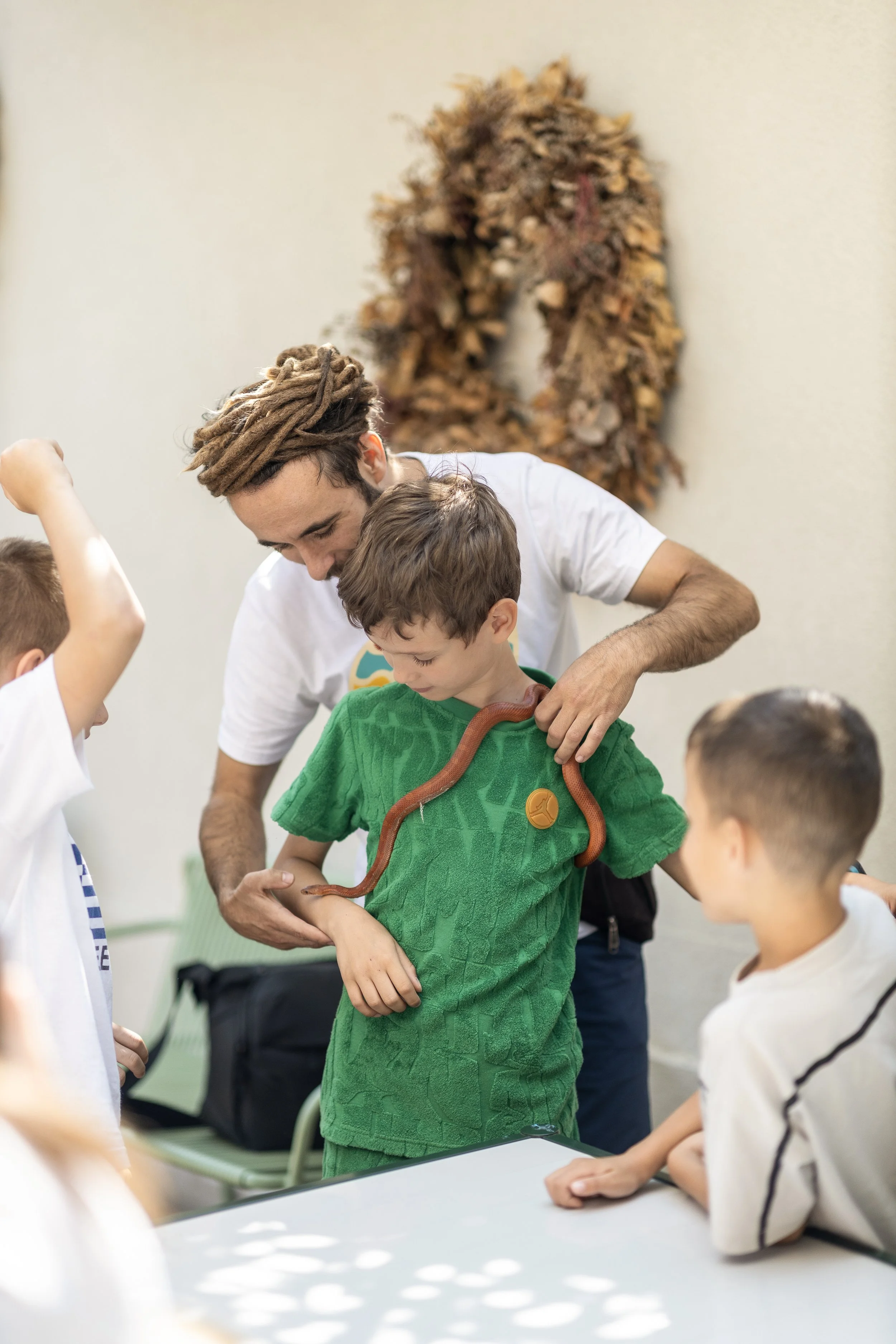 A group of children and a young man with dreadlocks looking at a brown snake on a child's arm during a presentation or educational demonstration in an indoor setting.