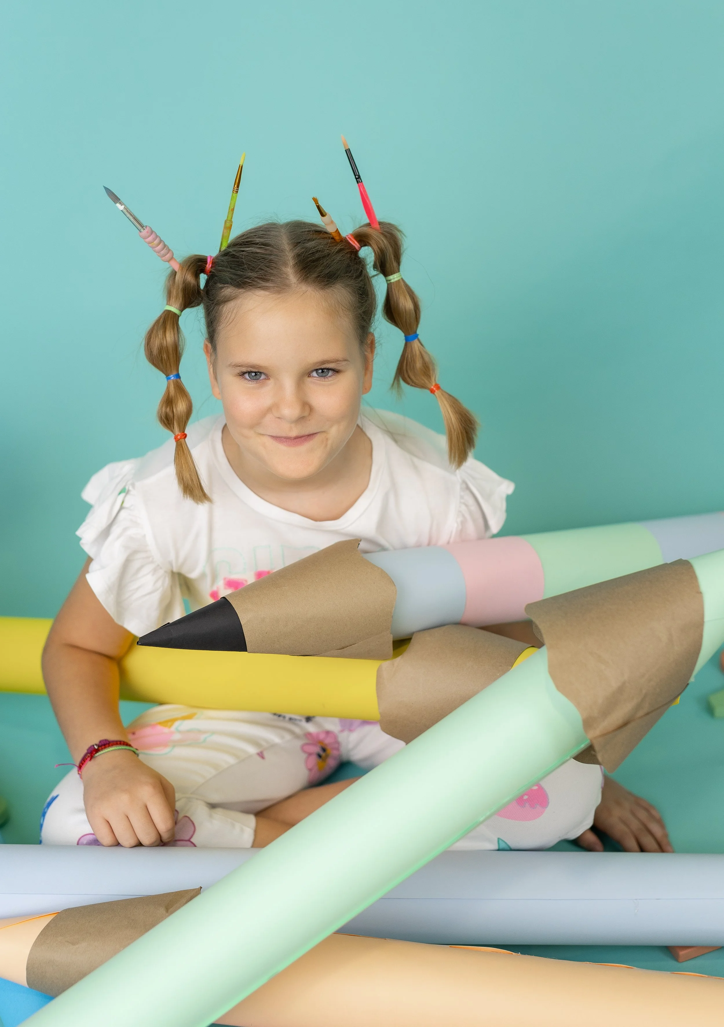 A young girl with braided hair and colorful hair ties, smiling at the camera, surrounded by rolled-up pastel-colored papers and brown craft paper, against a teal background.