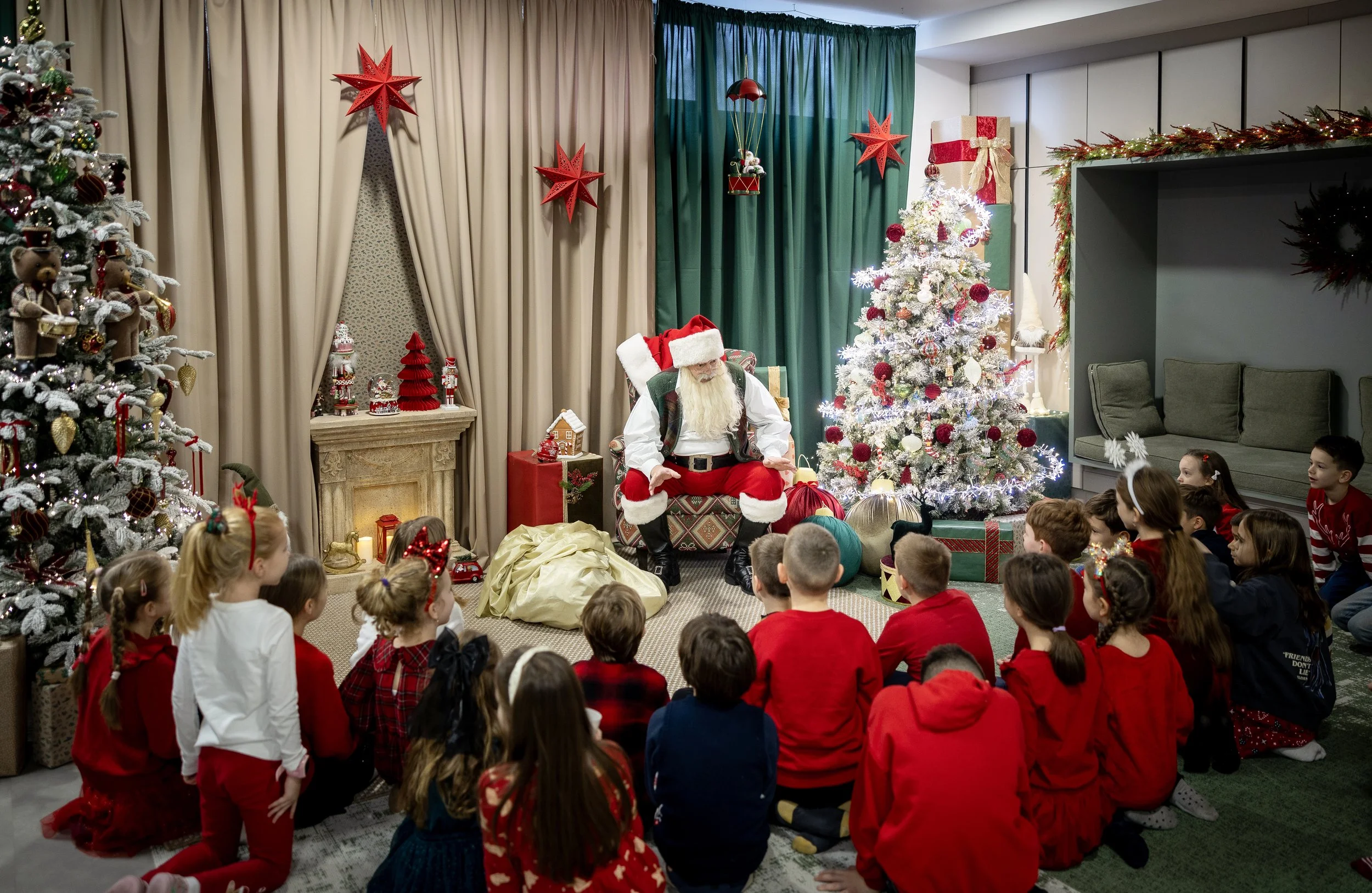 Children gathered around a Christmas tree watching Santa Claus sitting on a decorated chair in a festive room with Christmas decorations, including red stars, ornaments, and wrapped presents.
