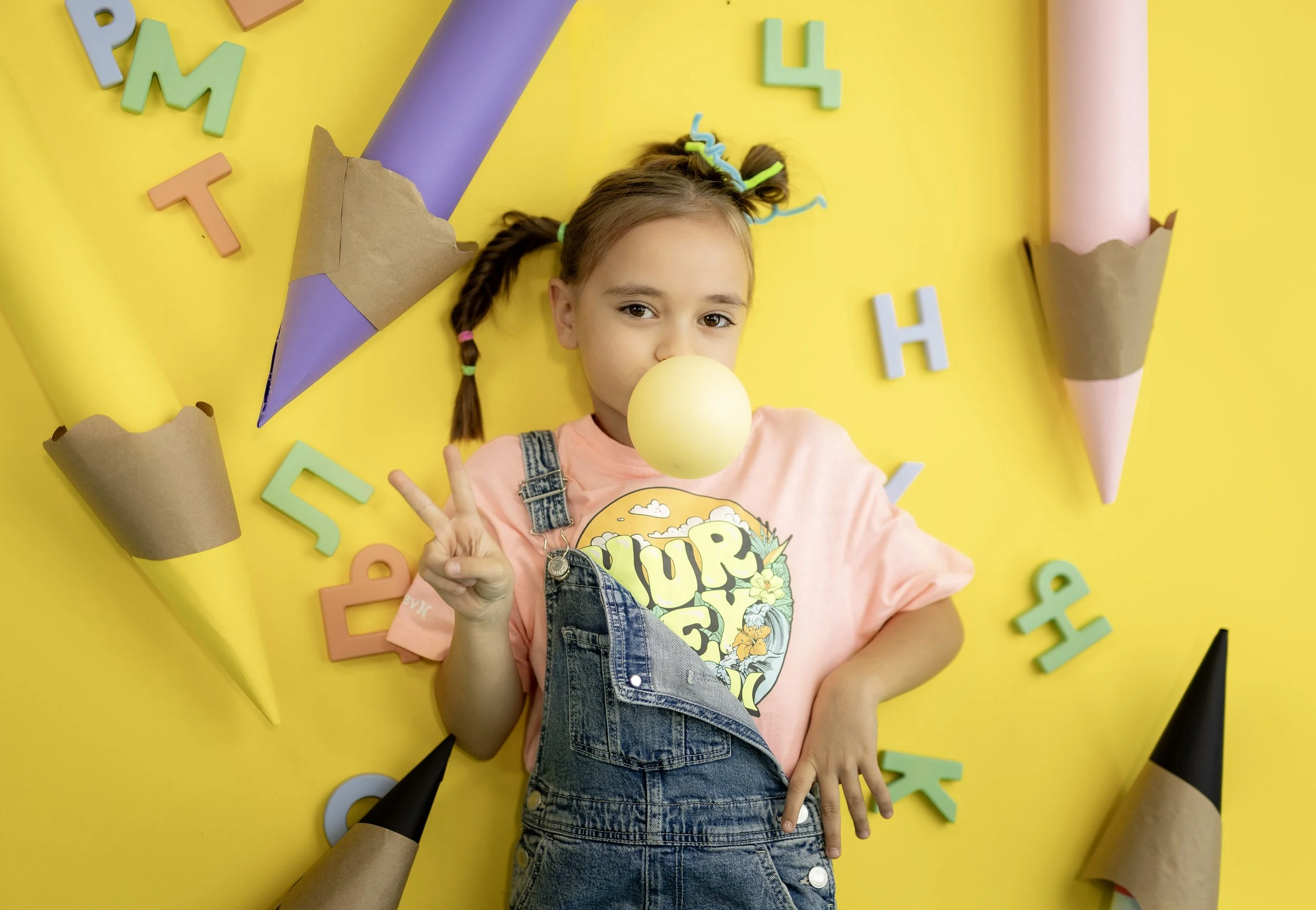 A young girl with pigtails holding yellow foam ball, lying on yellow background decorated with colorful foam letters and paper cones.