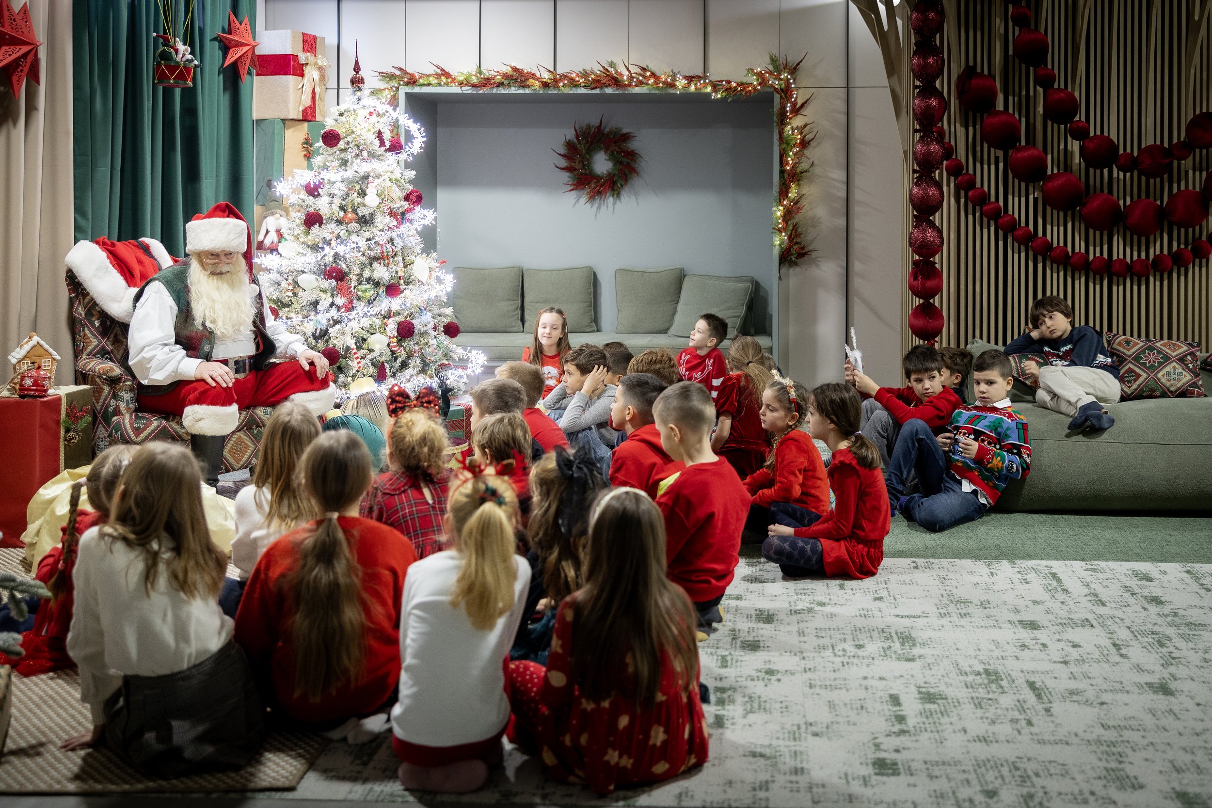 Children seated on the floor watching Santa Claus sitting nearby during a Christmas event. The room is decorated with a Christmas tree, wreath, and ornaments.