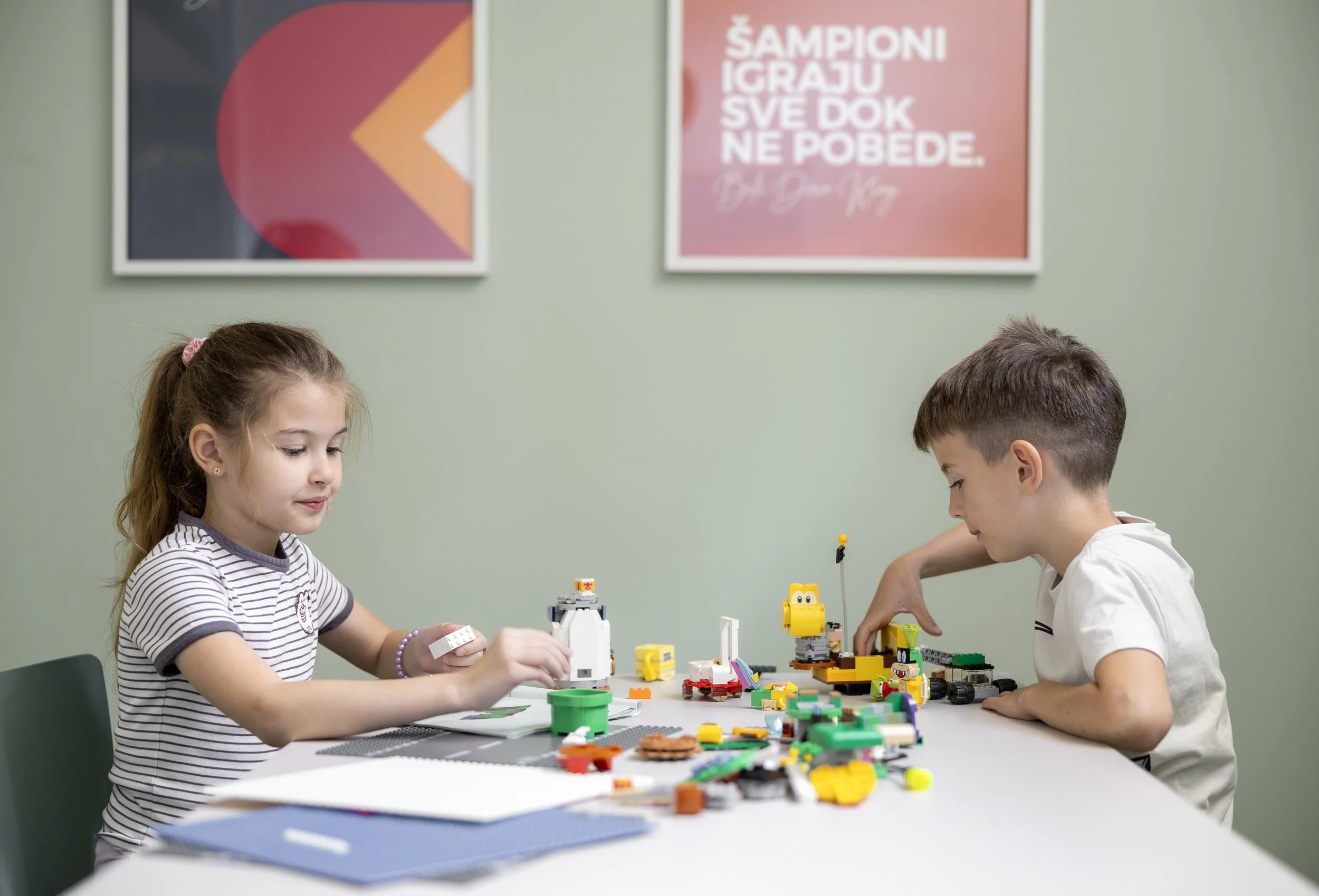 Two children, a girl and a boy, playing with colorful LEGO bricks on a white table in a classroom with light green walls. There are framed posters on the wall behind them, one displaying a quote in a foreign language.