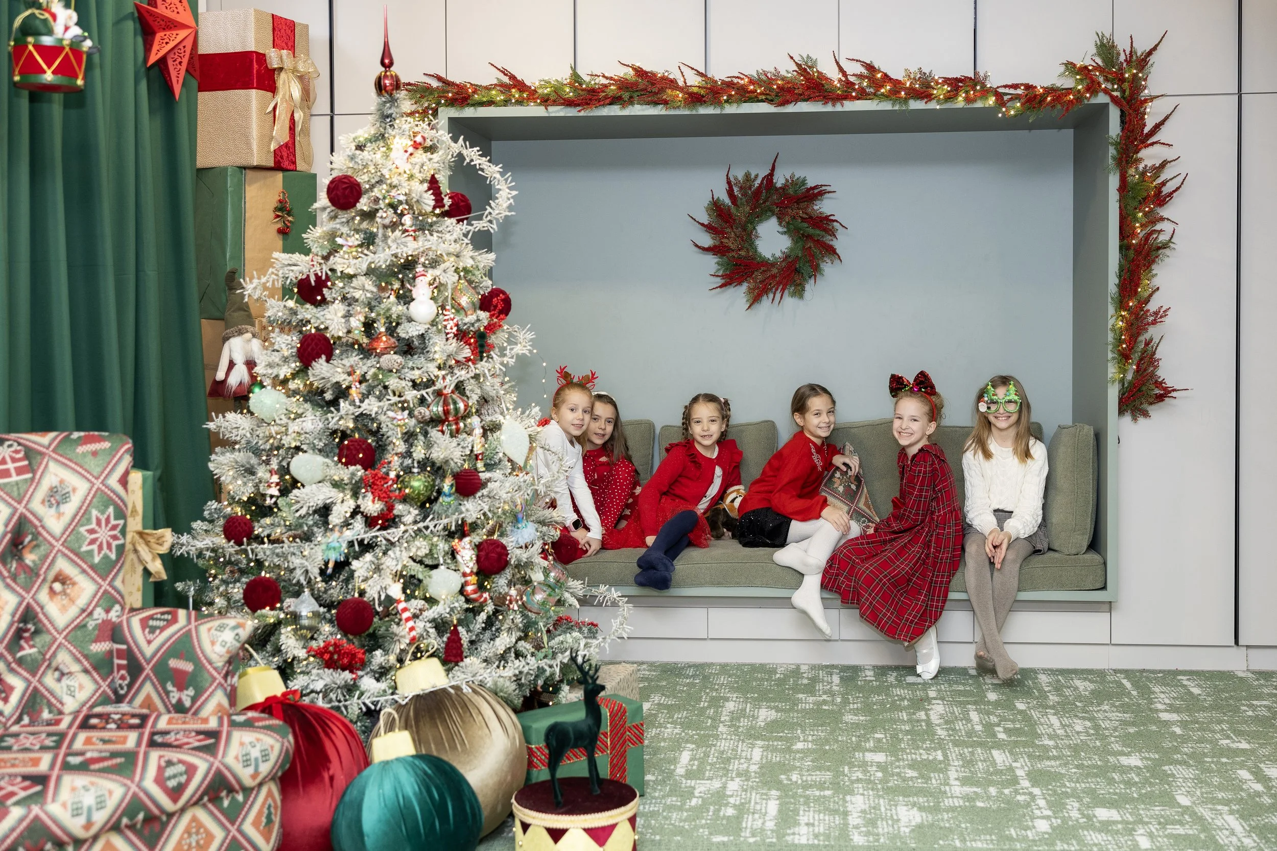 A group of seven young girls sitting on a couch in front of a decorated Christmas tree with wrapped presents nearby. The girls are dressed in festive clothing and some wear holiday accessories, smiling and posing for the photo.