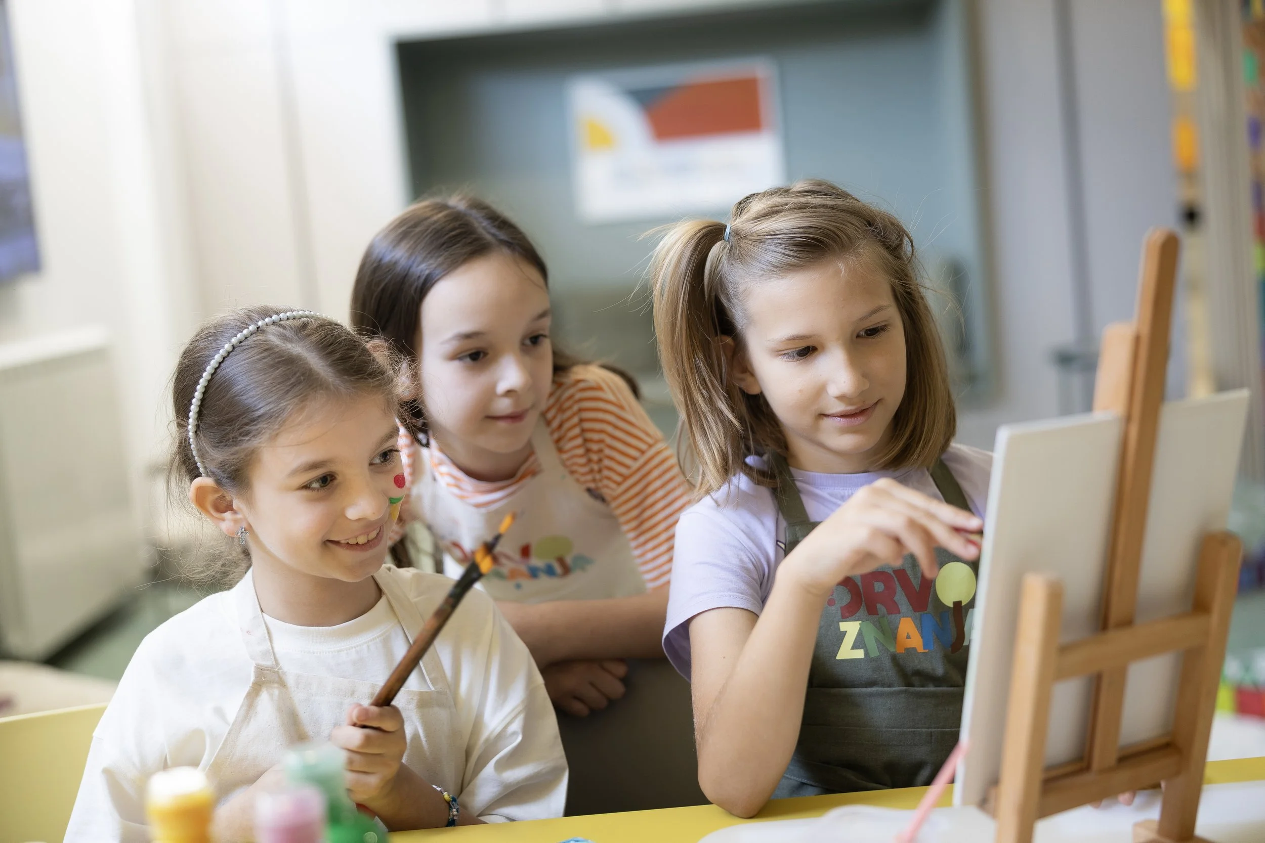 Four young girls gathered around an easel, painting on a canvas in an art classroom.
