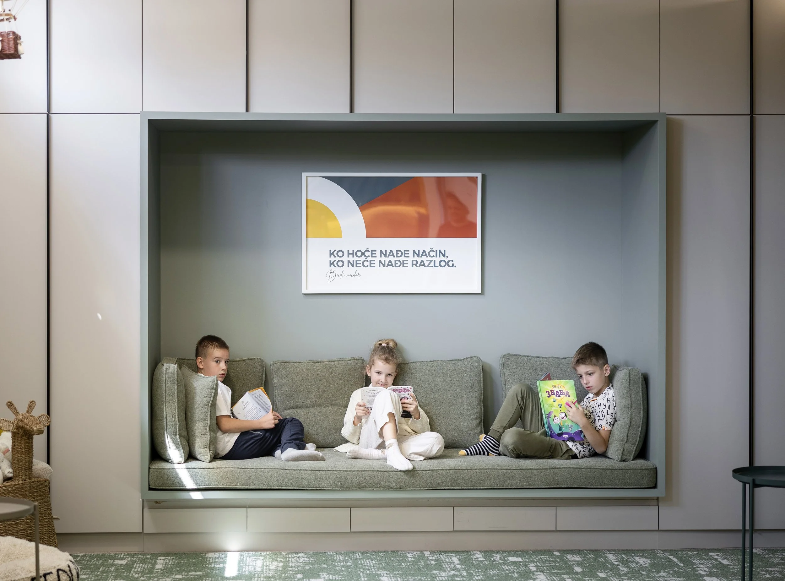 Four children sitting on a cushioned bench inside a modern, minimalist room. They are reading books, and each appears engaged with their reading material. The wall behind them features a framedabstract art print with shapes and colors. There is also a small table and a wicker basket with a plush toy on it in the room.