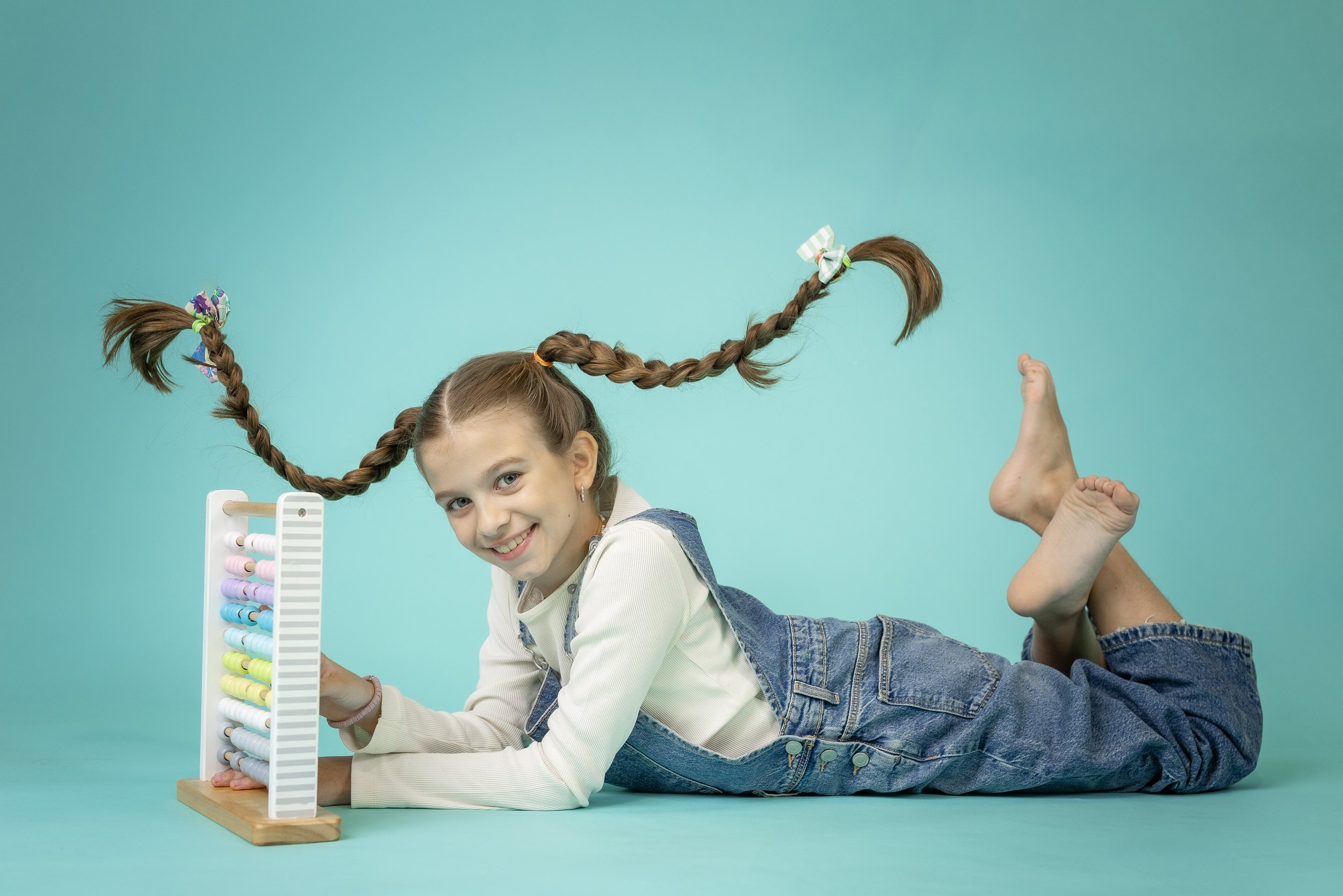 A young girl with braided hair and face paint, lying on a turquoise surface, smiling at the camera, with a colorful abacus toy in front of her and a person’s foot in the background.