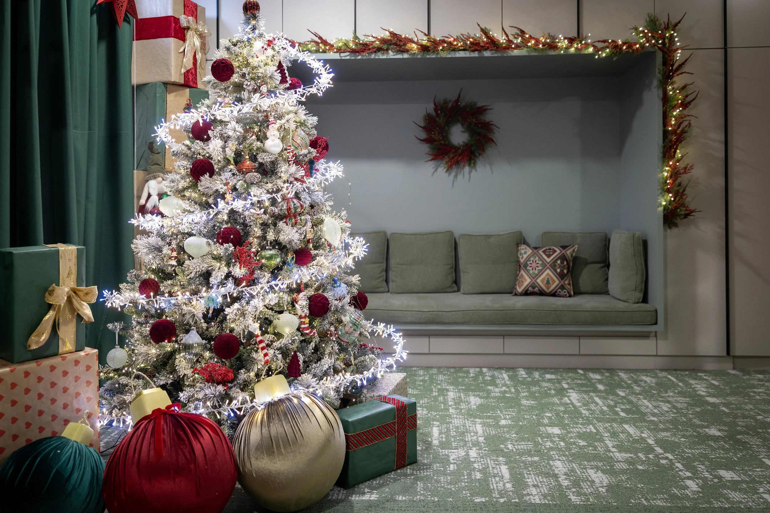 Christmas decorated room featuring a white Christmas tree with red, gold, and white ornaments and lights, with wrapped gifts underneath, and a gray cushioned window seat with patterned pillows, framed by red and green garland and wreaths.