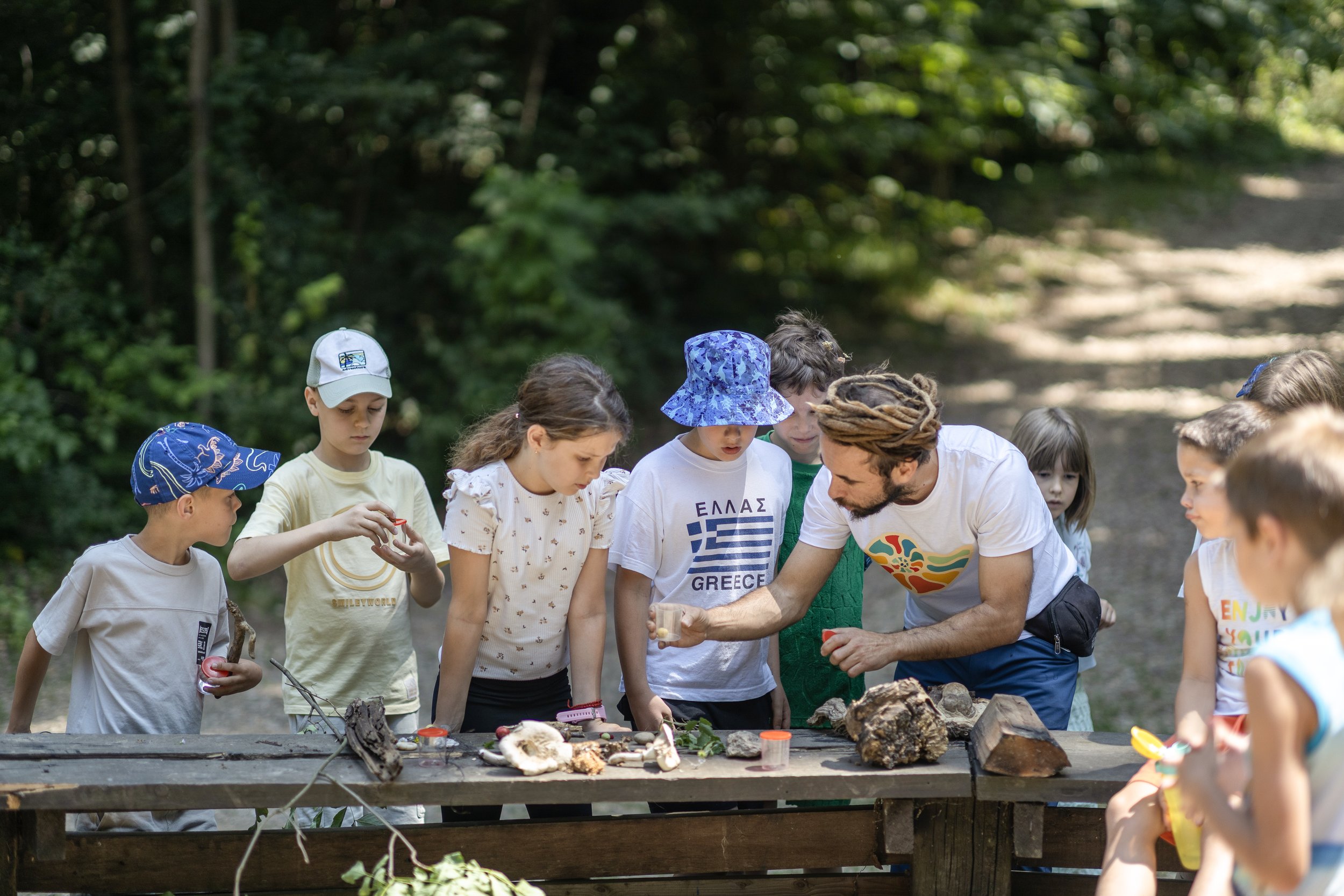Group of children and an adult gathered around a wooden table outdoors, examining objects on the table, with dense green trees in the background.