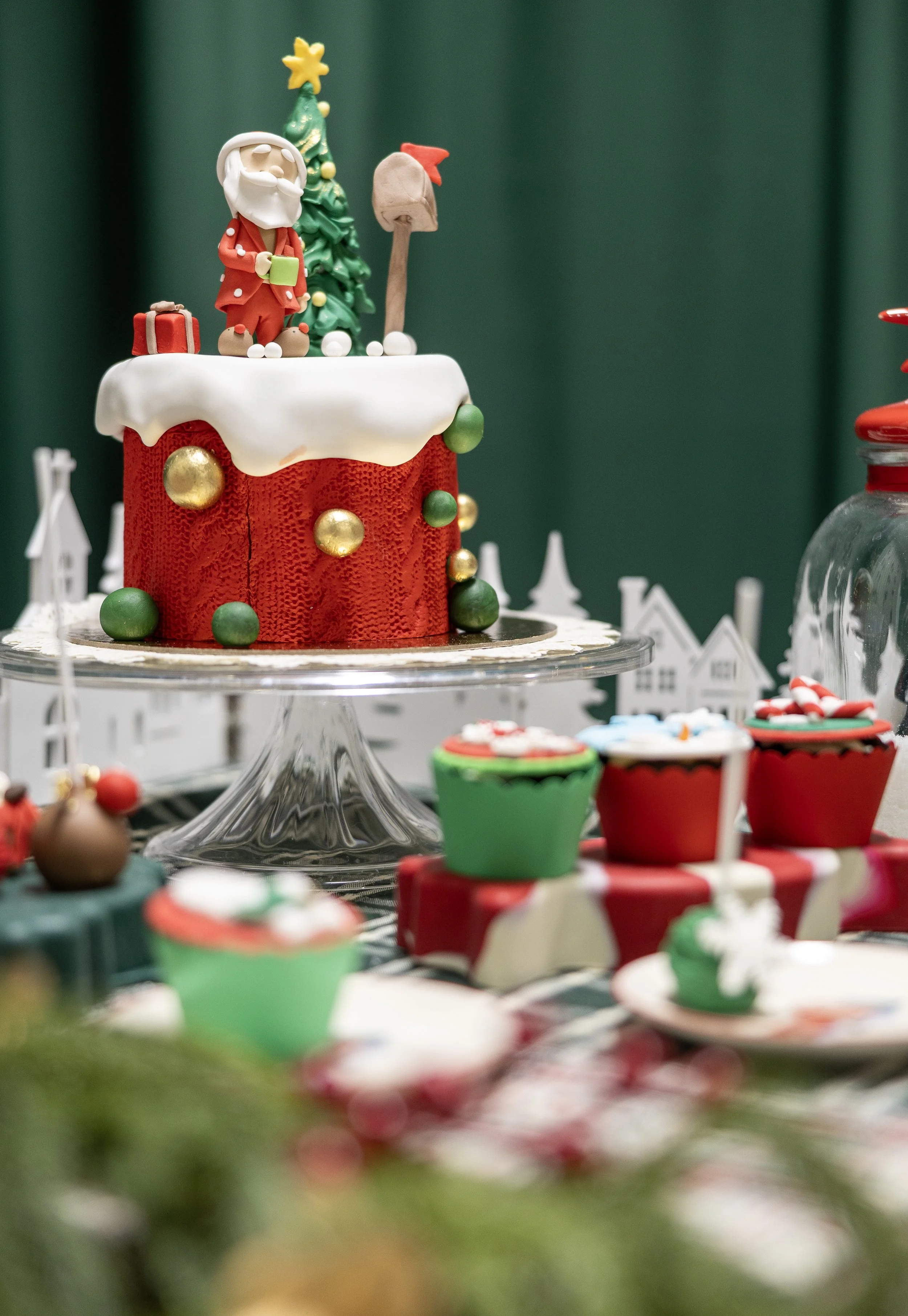 A Christmas-themed cake with Santa Claus, a decorated Christmas tree, and a candy cane on top, surrounded by holiday cupcakes and decorations.