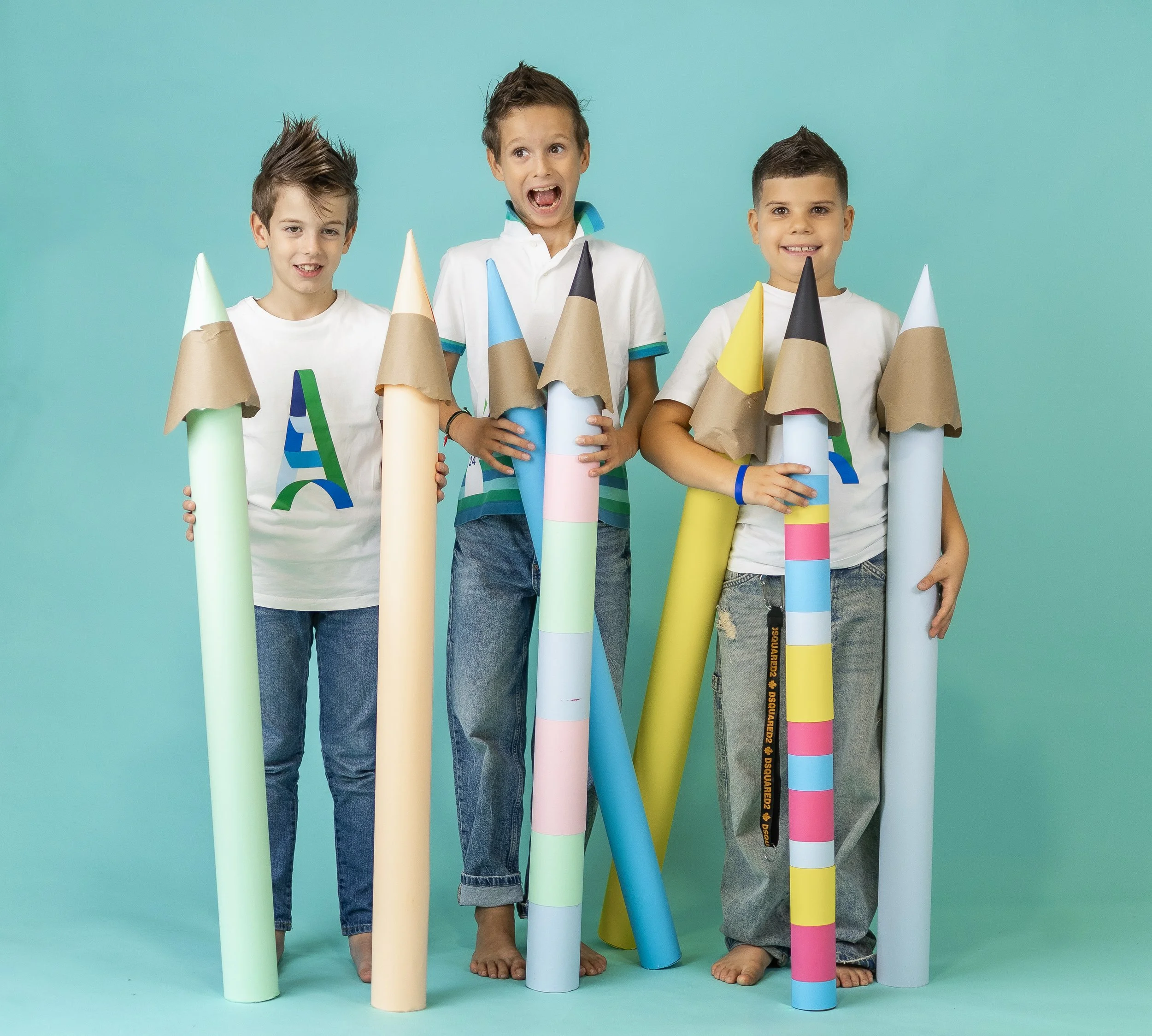 Three boys standing barefoot in front of a pastel blue background, holding large rolled paper objects decorated with colorful tape, creating a playful and vibrant scene.