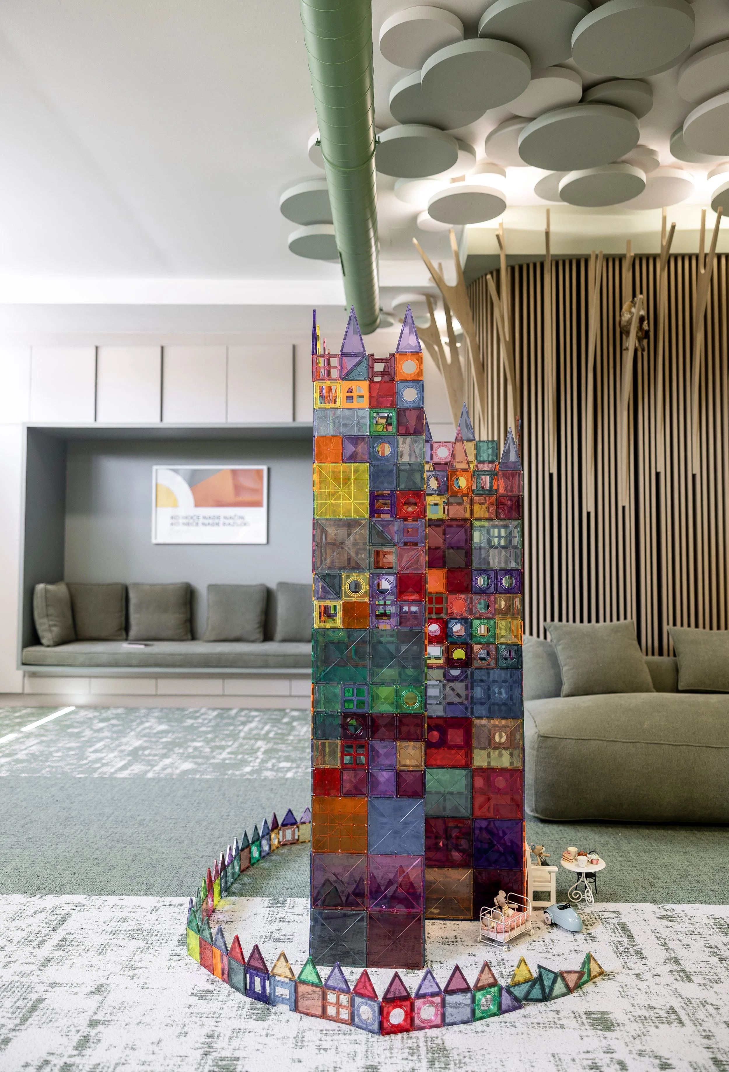 Colorful magnetic building blocks arranged to resemble a castle or skyscraper, set on a carpeted floor in a modern lounge area with gray couches and wooden wall decor.
