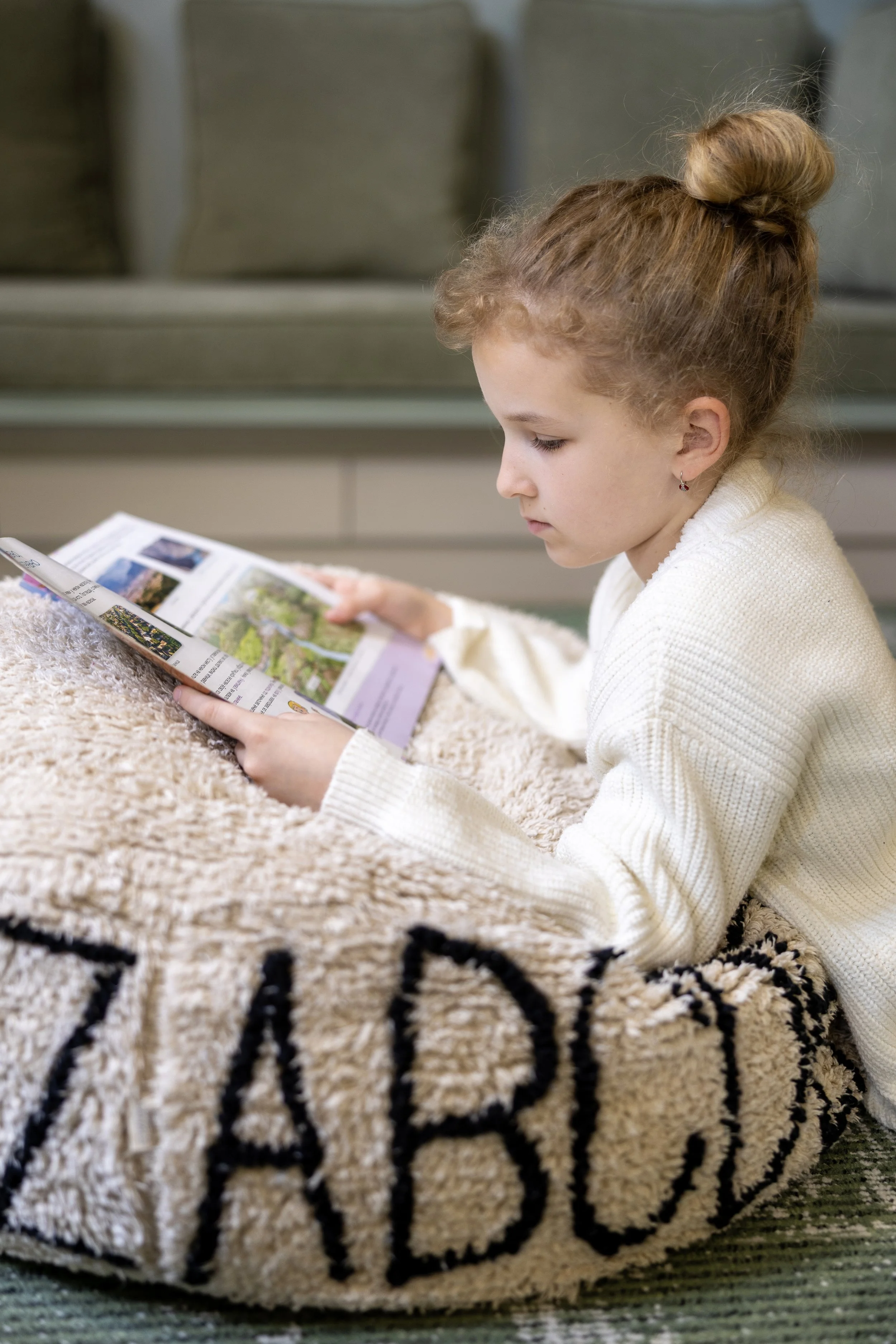 A young girl with curly red hair in a bun is sitting on a beige fuzzy blanket and reading a magazine, with a gray couch in the background.