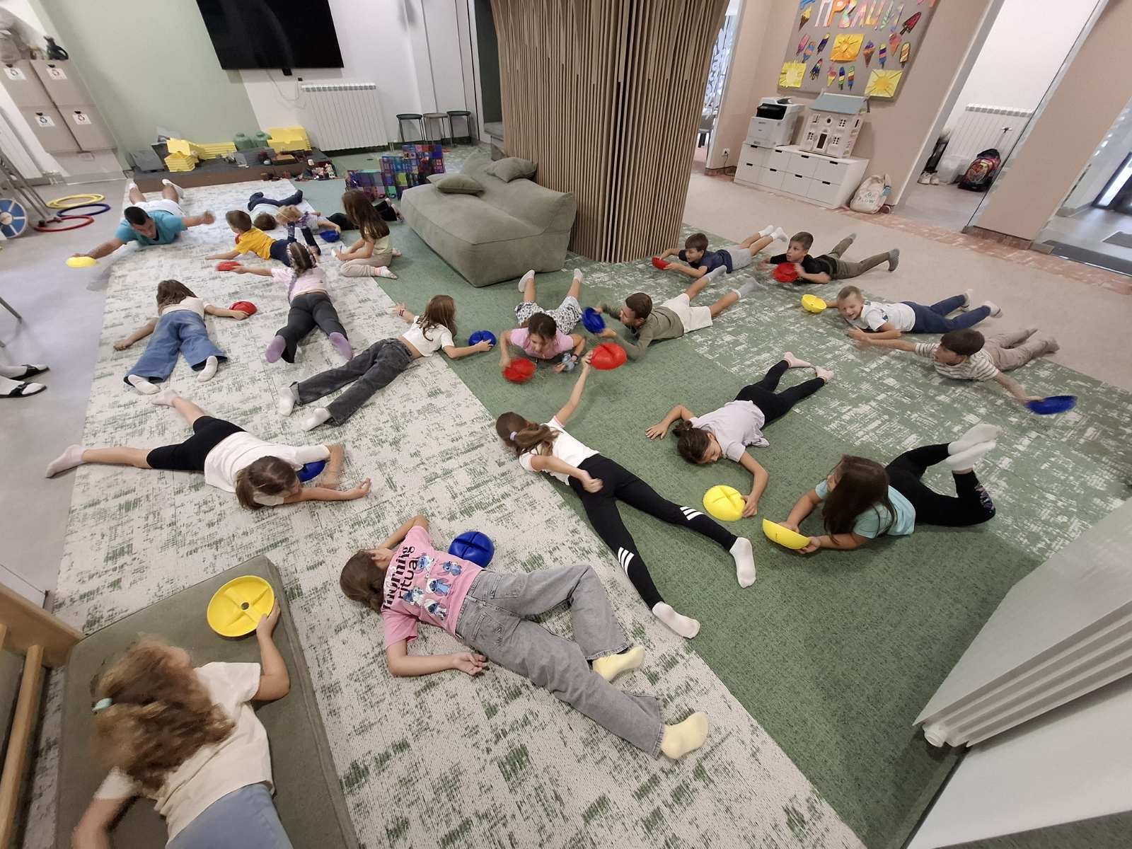 Children lying on the floor in a large room, engaging in a playful activity with colorful plastic turtles.