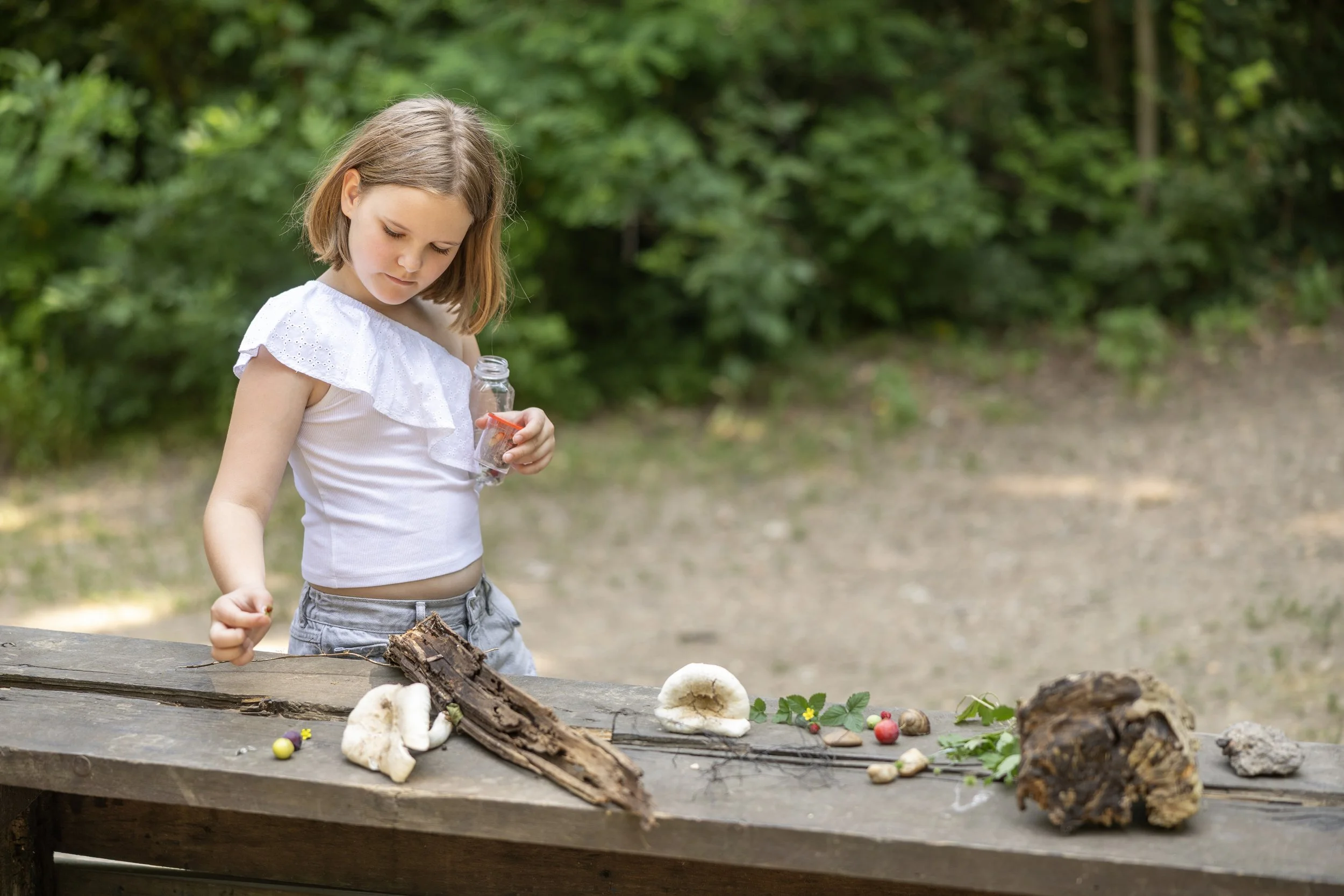 A young girl with shoulder-length brown hair wearing a white top and gray shorts stands outdoors near a wooden table with various natural objects like mushrooms, bark, stones, and small decorated eggs, in front of a green leafy background.