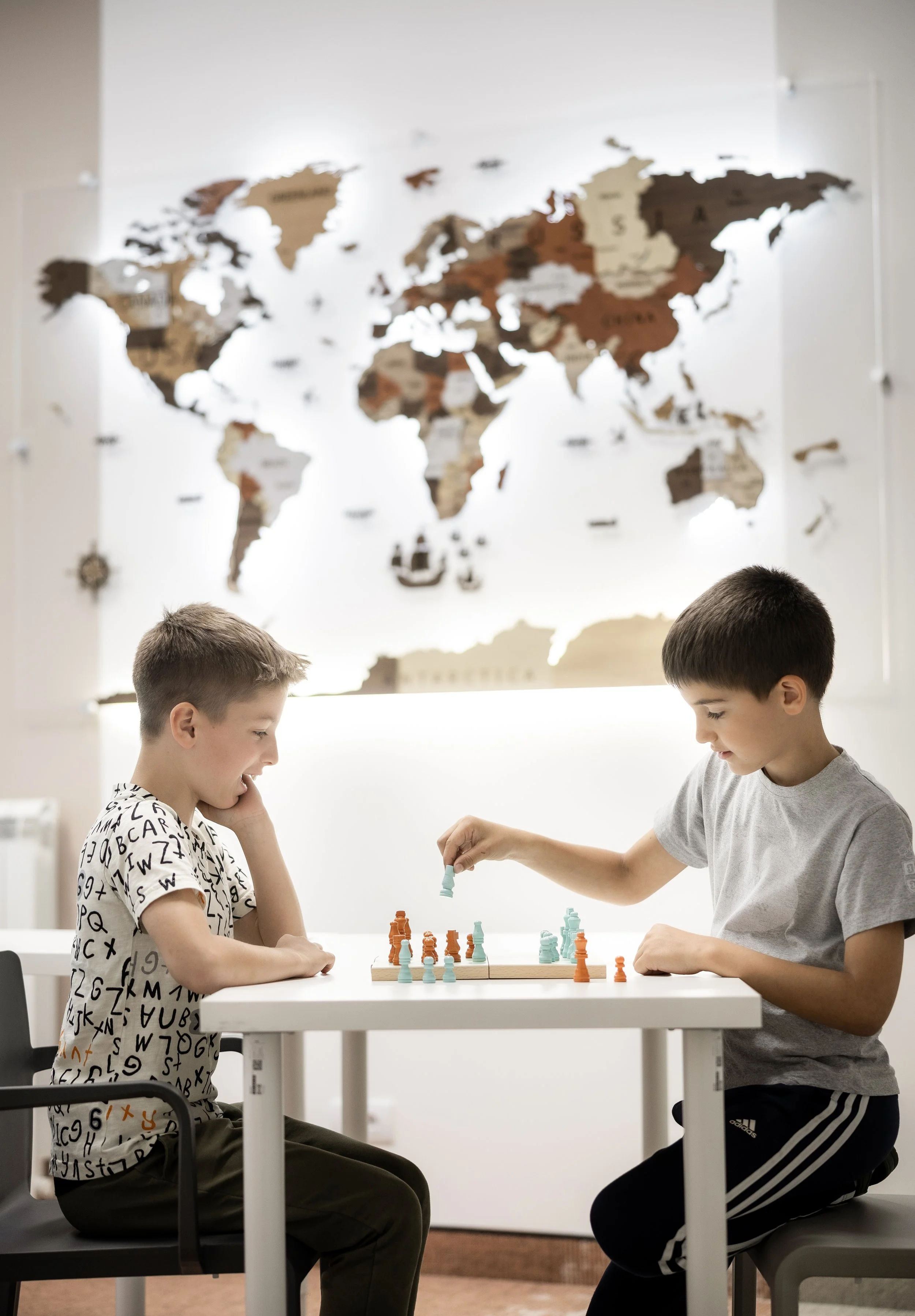 Two boys playing chess at a white table in a room with a world map on the wall.