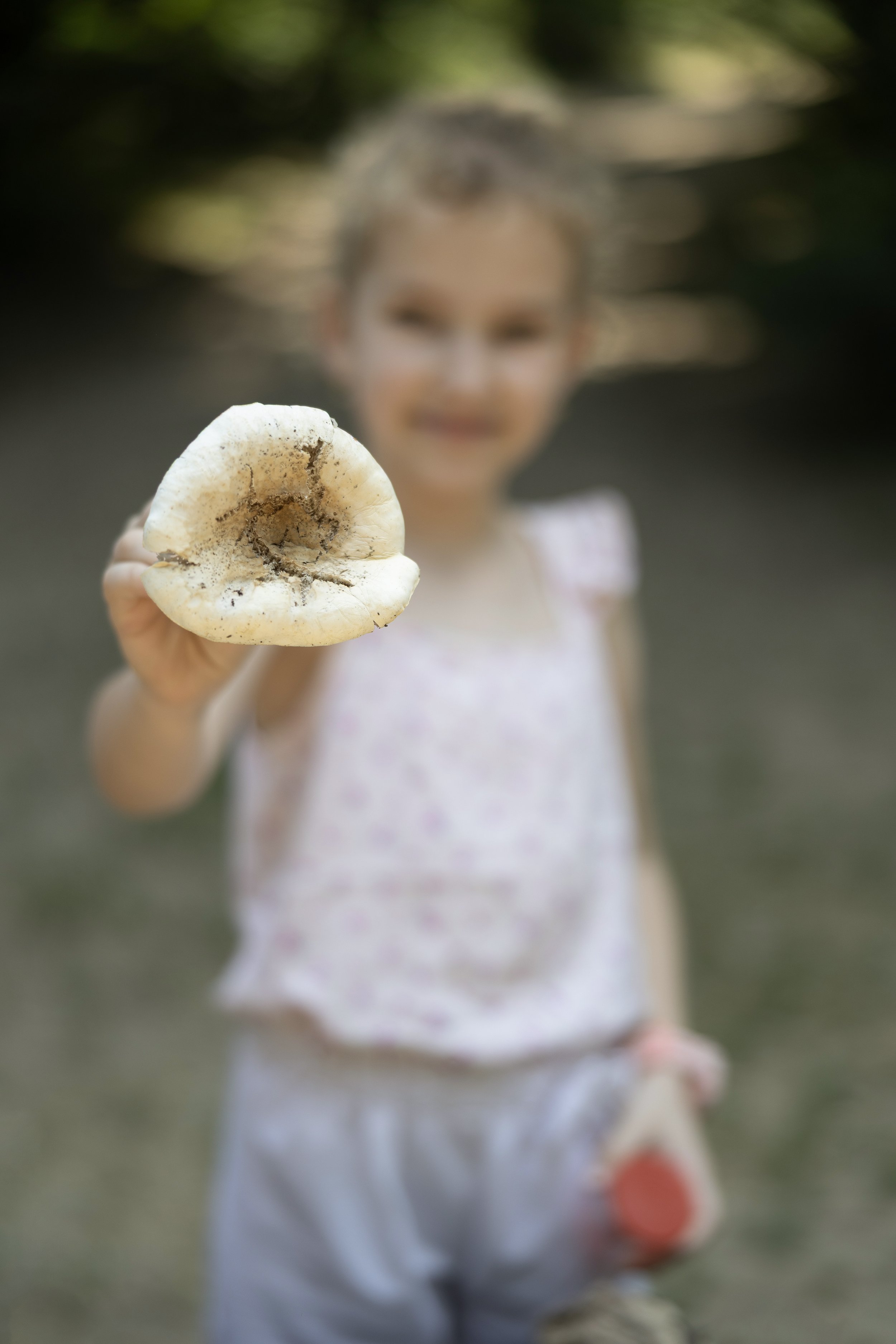 A young girl holding a large mushroom towards the camera with a blurred background of green trees.