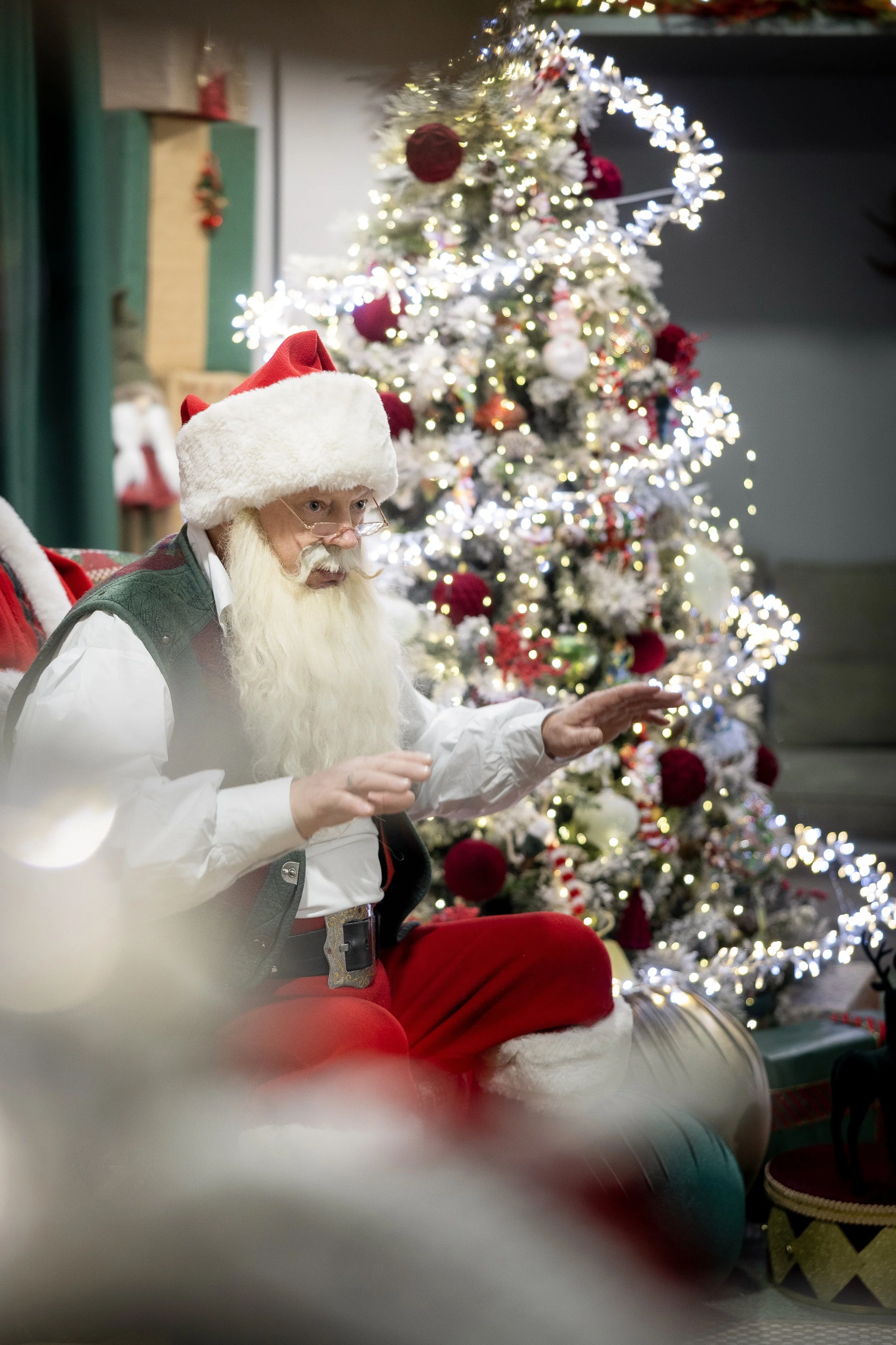 A person dressed as Santa Claus with a white beard, wearing glasses, a Santa hat, a white shirt, and red pants, sitting in front of a decorated Christmas tree with white and red ornaments and lights, within a cozy room.