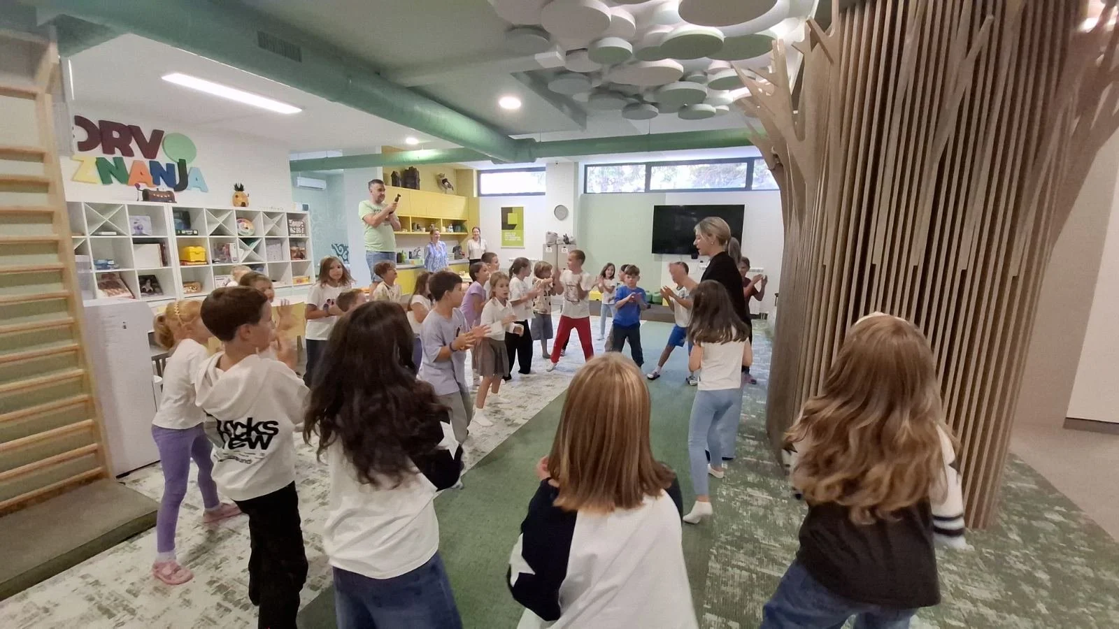 Children participating in a group activity led by a woman in an indoor playroom, with shelves filled with toys and books in the background.