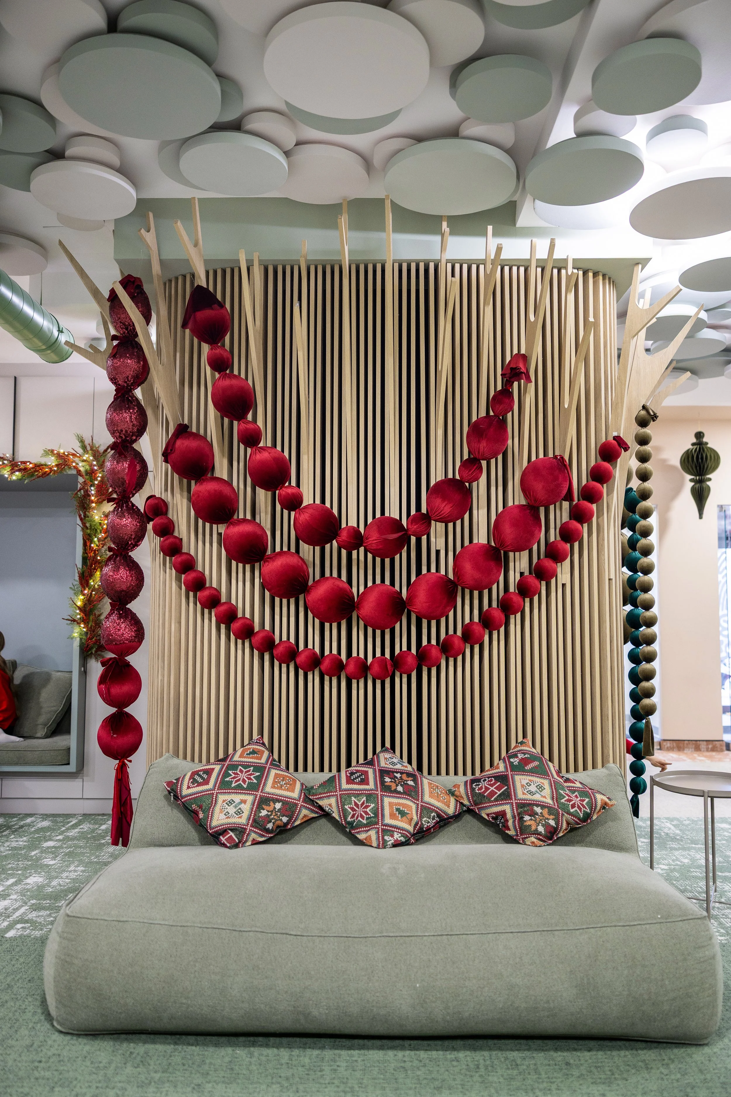 A modern sitting area with a light green sofa, four decorative pillows with geometric patterns, and Christmas decorations including a red garland, red ornaments, and a string of large beads. The background features a wooden slat wall, and the ceiling has a unique pattern of white and light green circular panels.