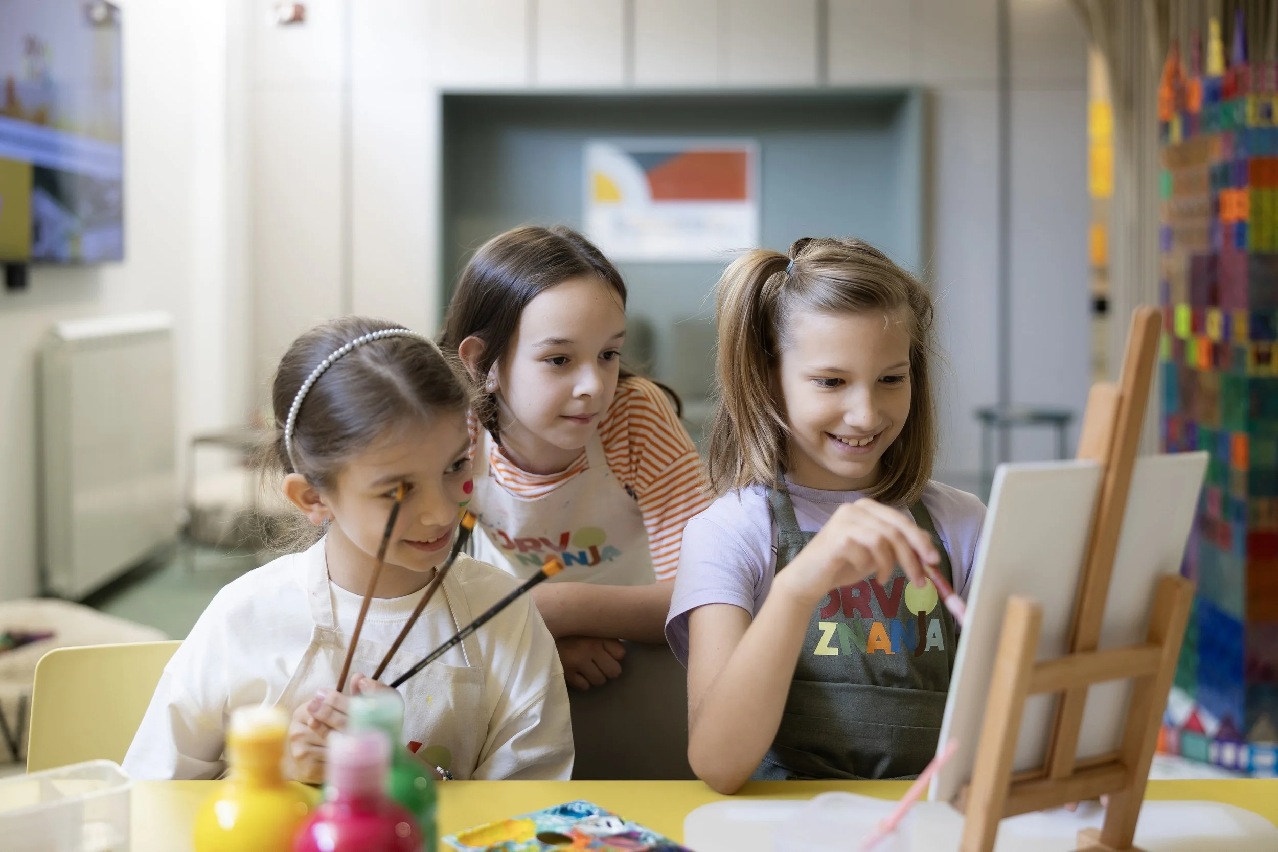 Four young girls painting in an art class, with colorful bottles of paint and an easel in front of them, focused on their artwork.
