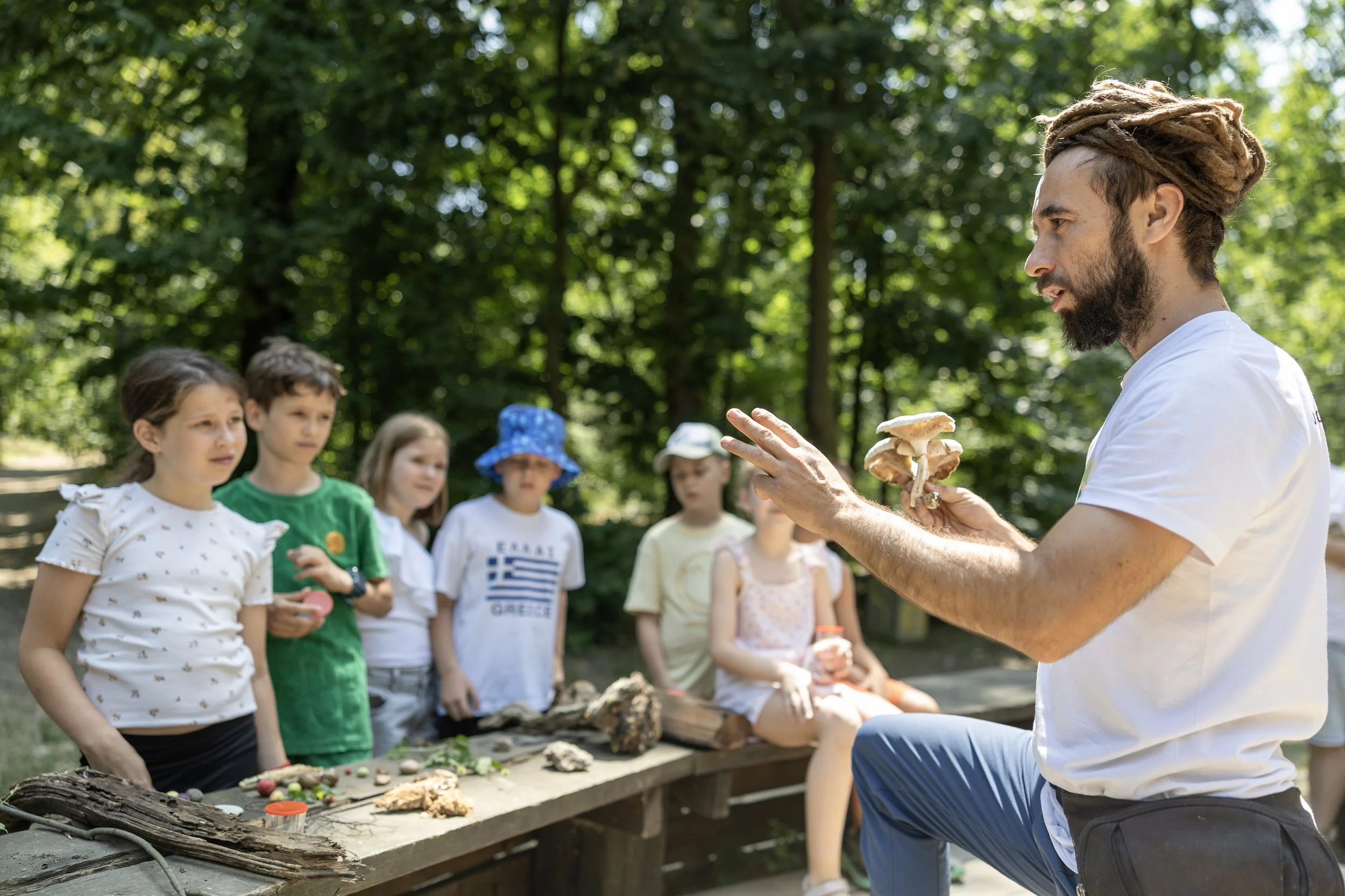 A man with dreadlocks and a beard demonstrates mushrooms to a group of children in a forest setting, with a table of natural objects and mushrooms.