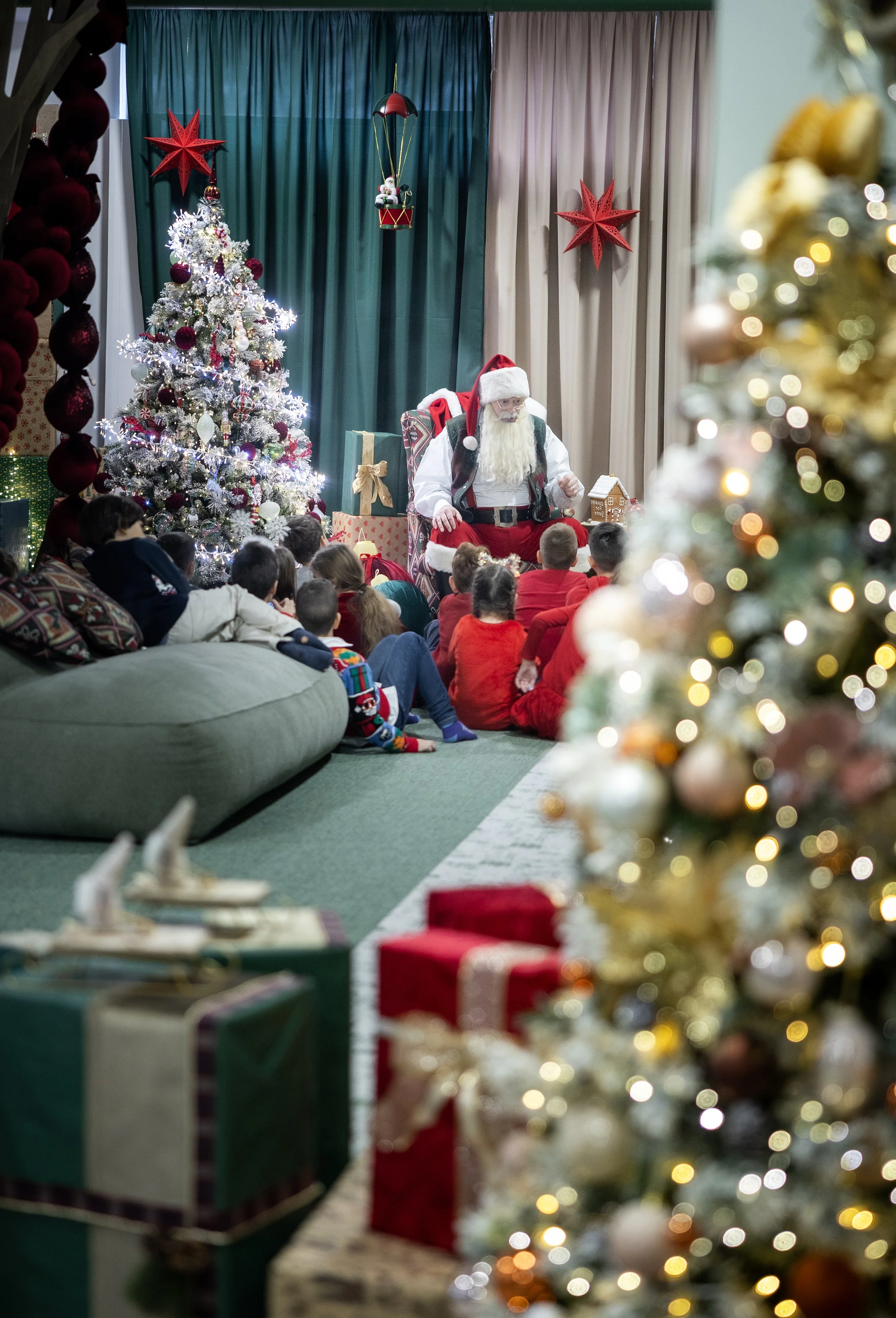 Santa Claus sitting on a chair with children gathered around during a Christmas celebration, decorated with a Christmas tree, presents, and holiday ornaments.