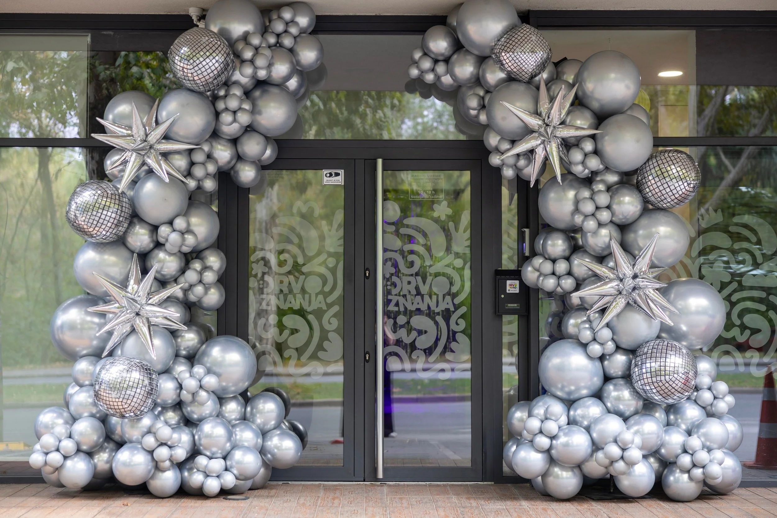 Entrance decorated with silver balloons, including star-shaped and disco ball-style balloons, surrounding glass doors with decorative patterns.