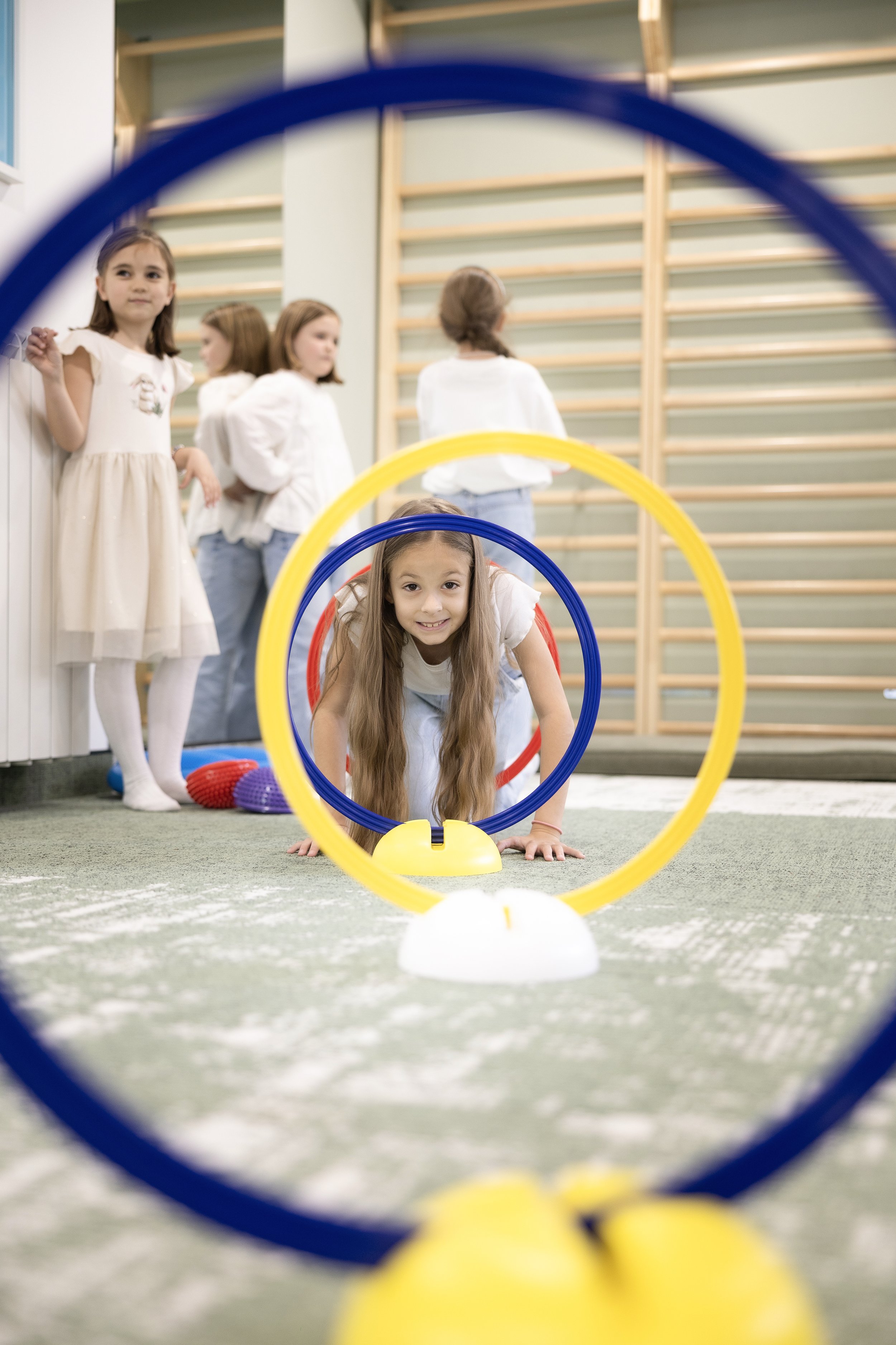 Young girl crawling through a yellow hoop during a physical activity in a gym, with other girls waiting in the background.