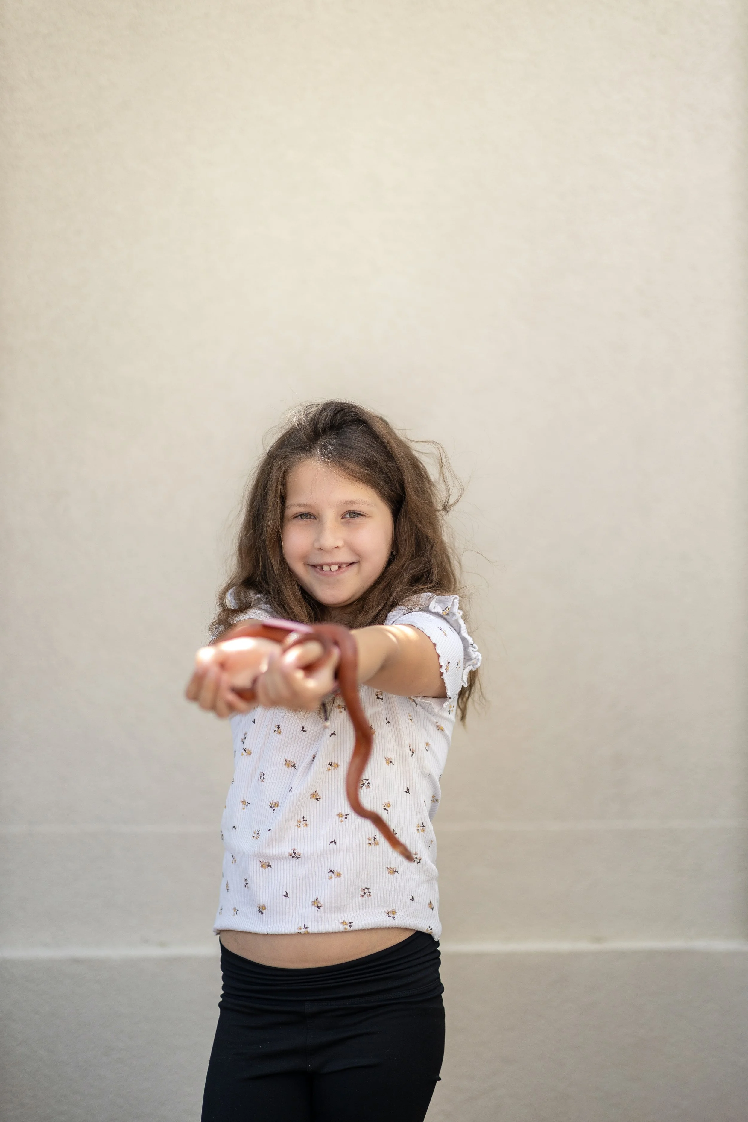 A young girl with long, wavy brown hair smiling and pointing a snake towards the camera. She is wearing a white, short-sleeved shirt with a small floral pattern and black pants, standing against a plain light-colored wall.