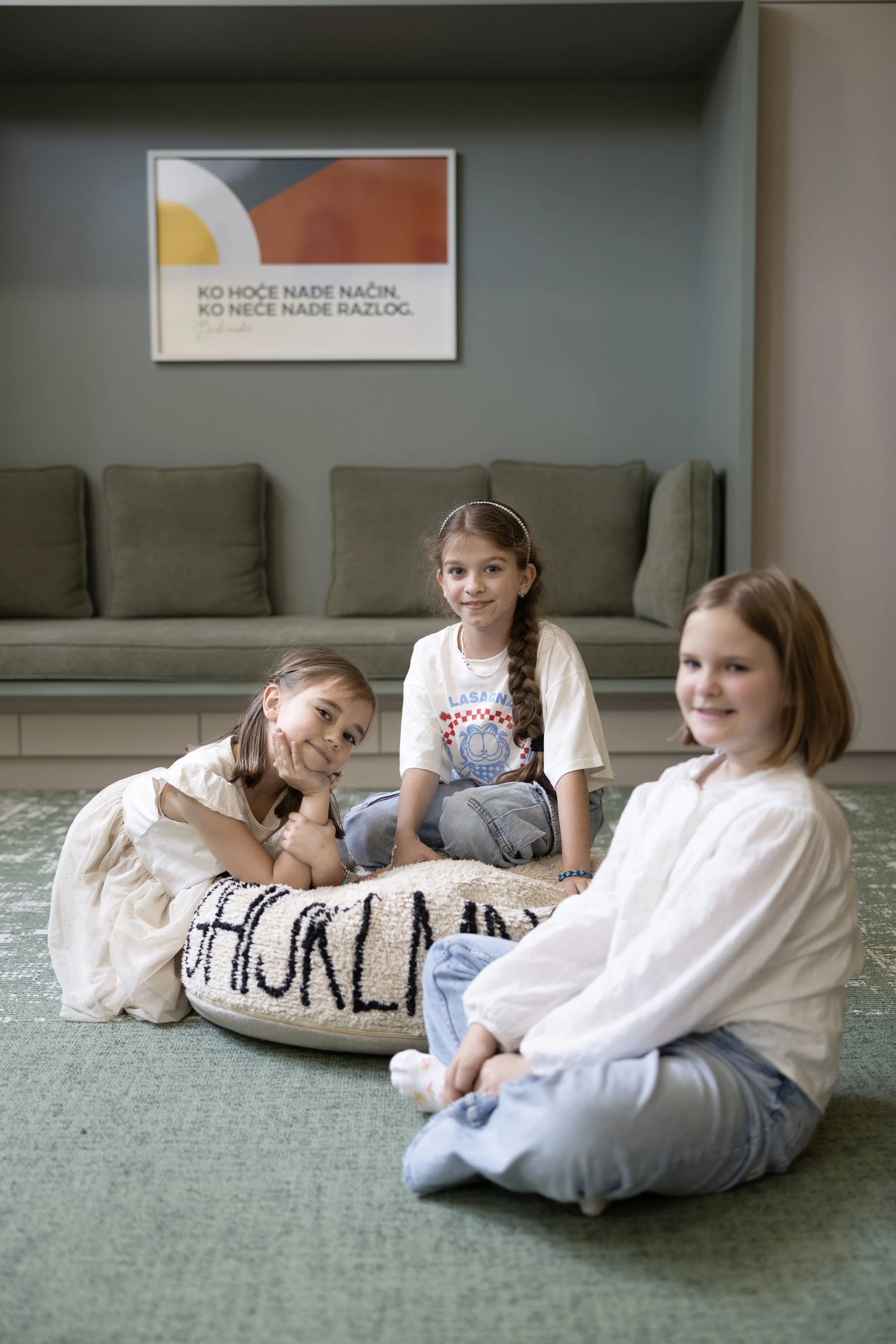 Four young girls sitting on a green carpet in a living room. Behind them, a gray sofa with cushions. A framed abstract art piece hangs on the wall with text beneath it. The girls are smiling and wearing casual clothes.