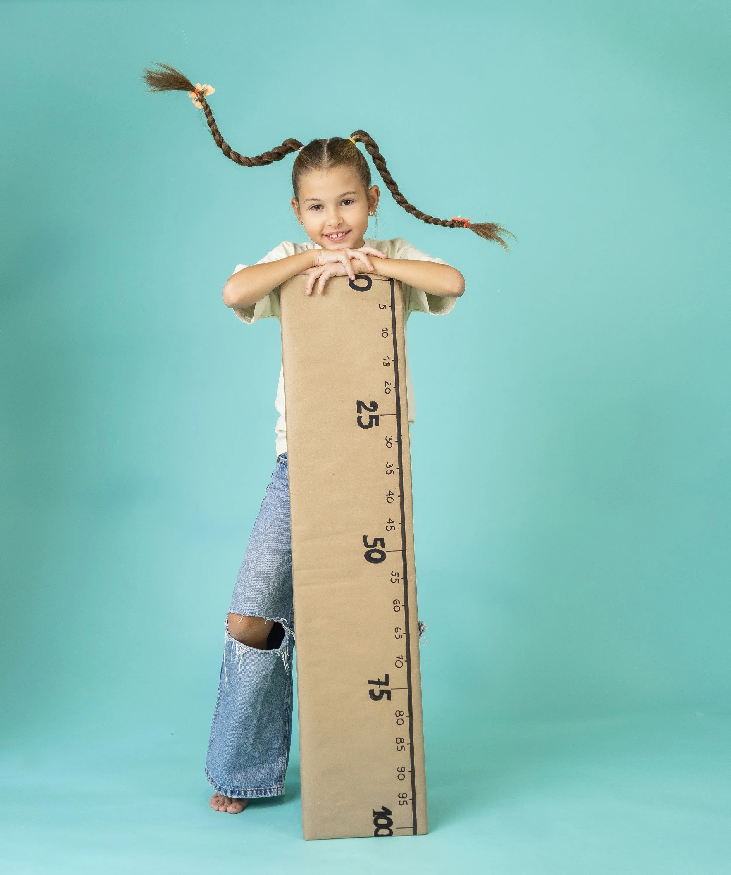 A young girl with braids, wearing a beige shirt and ripped jeans, leaning her arms on a large measuring stick that reaches over 100 centimeters, against a light blue background.