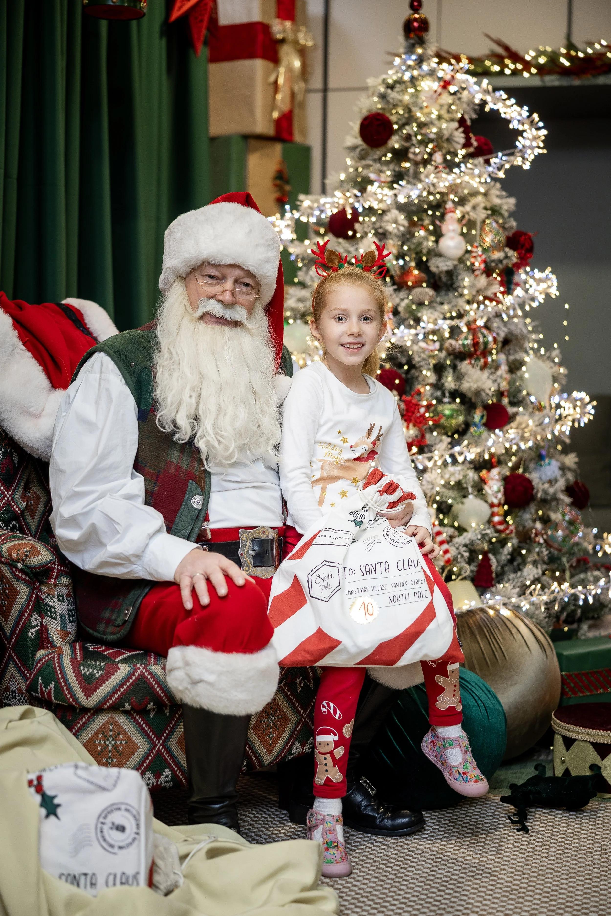A Christmas scene with a man dressed as Santa Claus and a girl sitting on Santa's lap. They are in a festive room with a decorated Christmas tree and wrapped presents. The girl is wearing Christmas-themed leggings and colorful shoes. Santa is wearing his traditional red suit with white trim, and the girl is holding a large, decorated pillow that looks like a wrapped gift with a mailing address to Santa Claus.