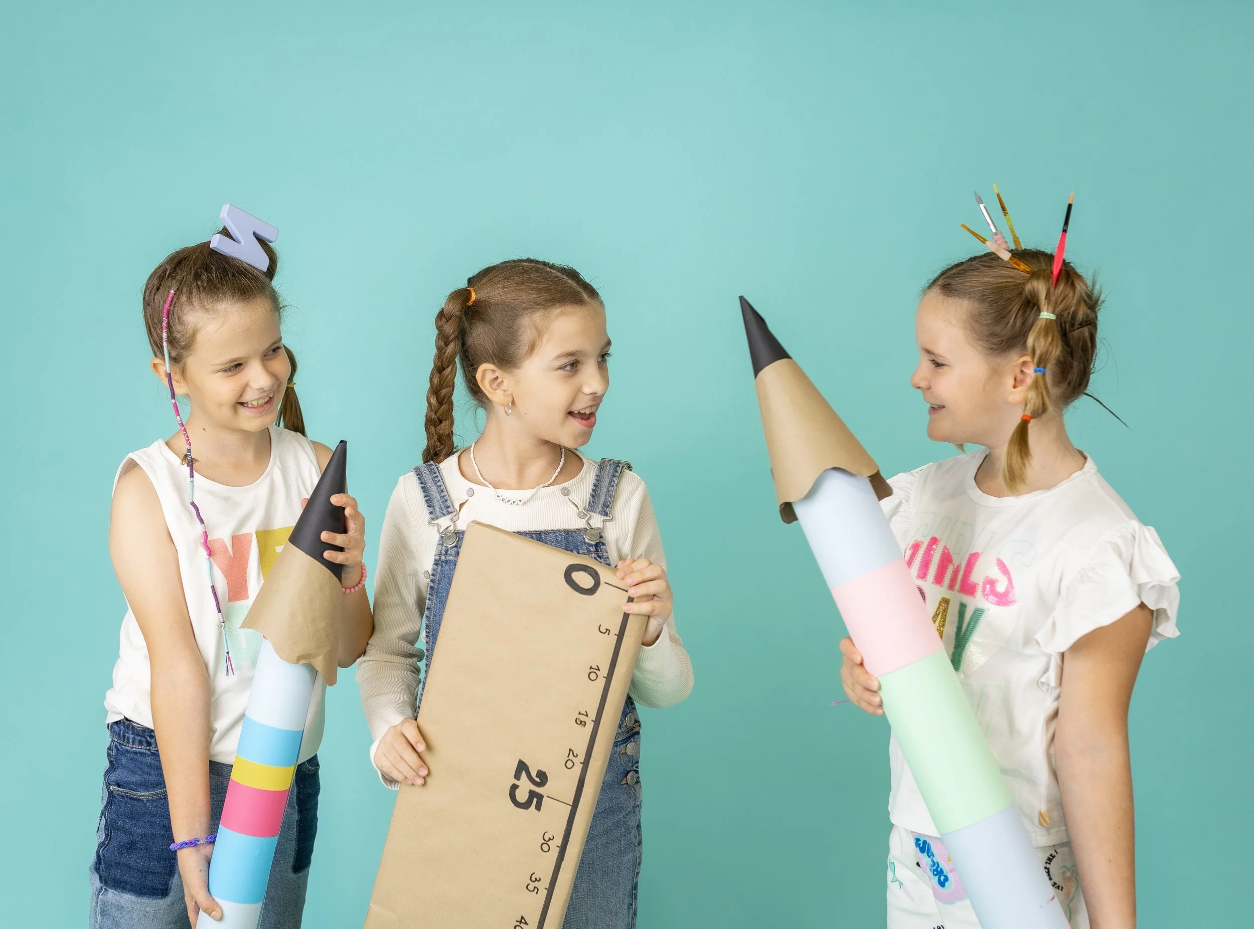 Three young girls smiling and wearing cardboard rocket props and headbands with colored pencils against a turquoise background.