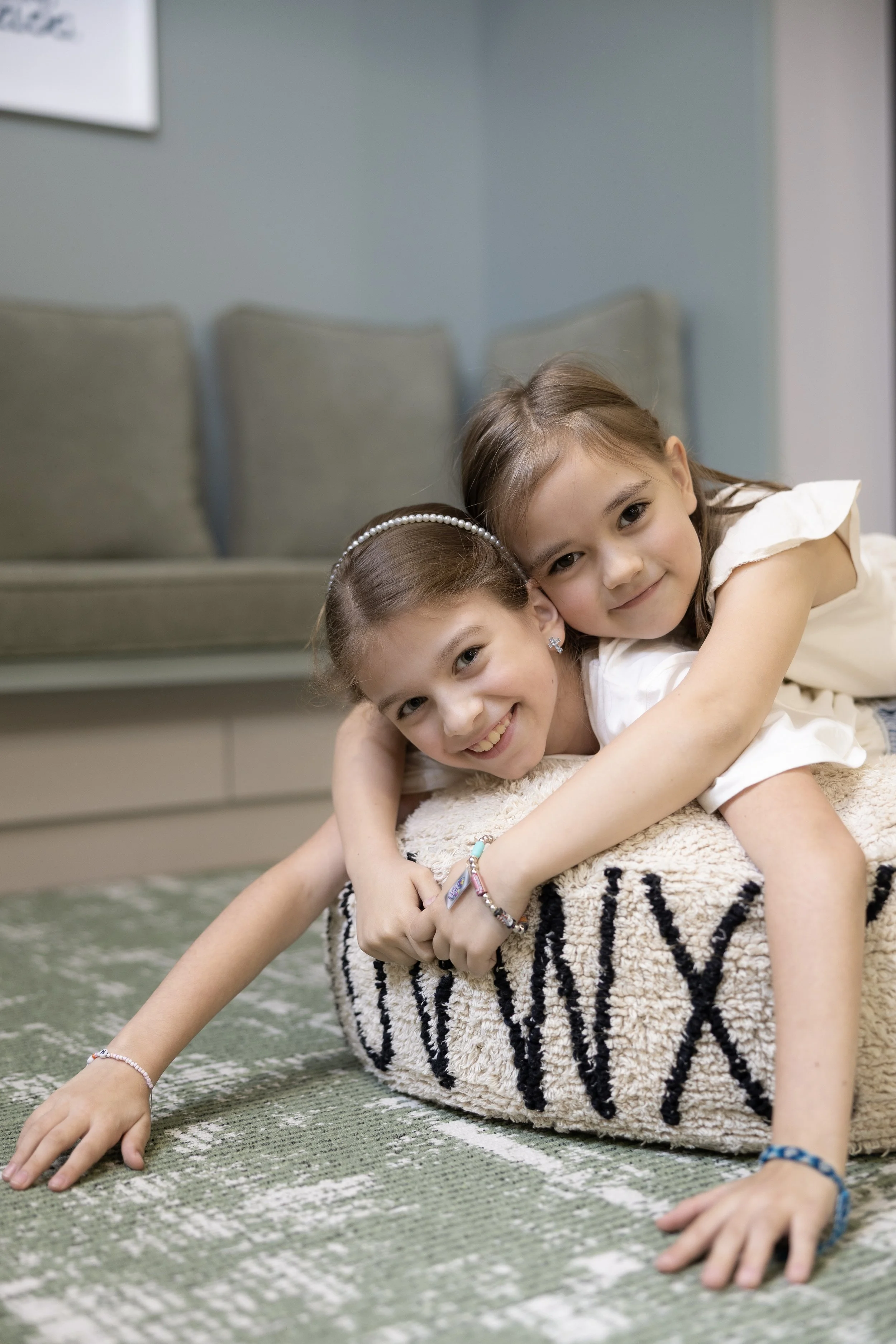 Two young girls lying on a green patterned rug, smiling and hugging, with a gray couch and light blue wall in the background.