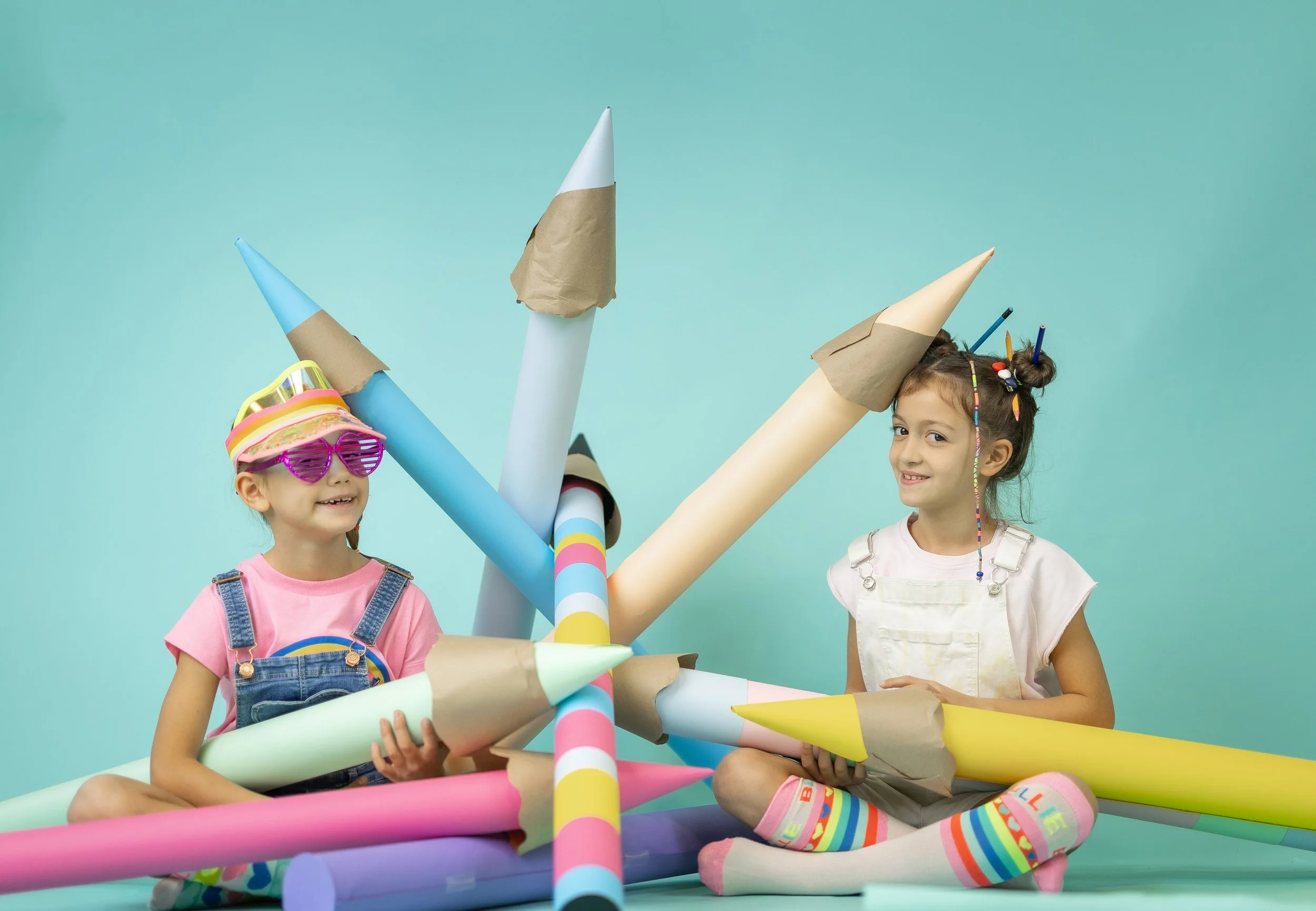 Two young girls playing with a colorful paper mache rocket ship against a bright blue background. One girl has a pink shirt, denim overalls, and sunglasses, while the other is in a white shirt and striped rainbow socks.