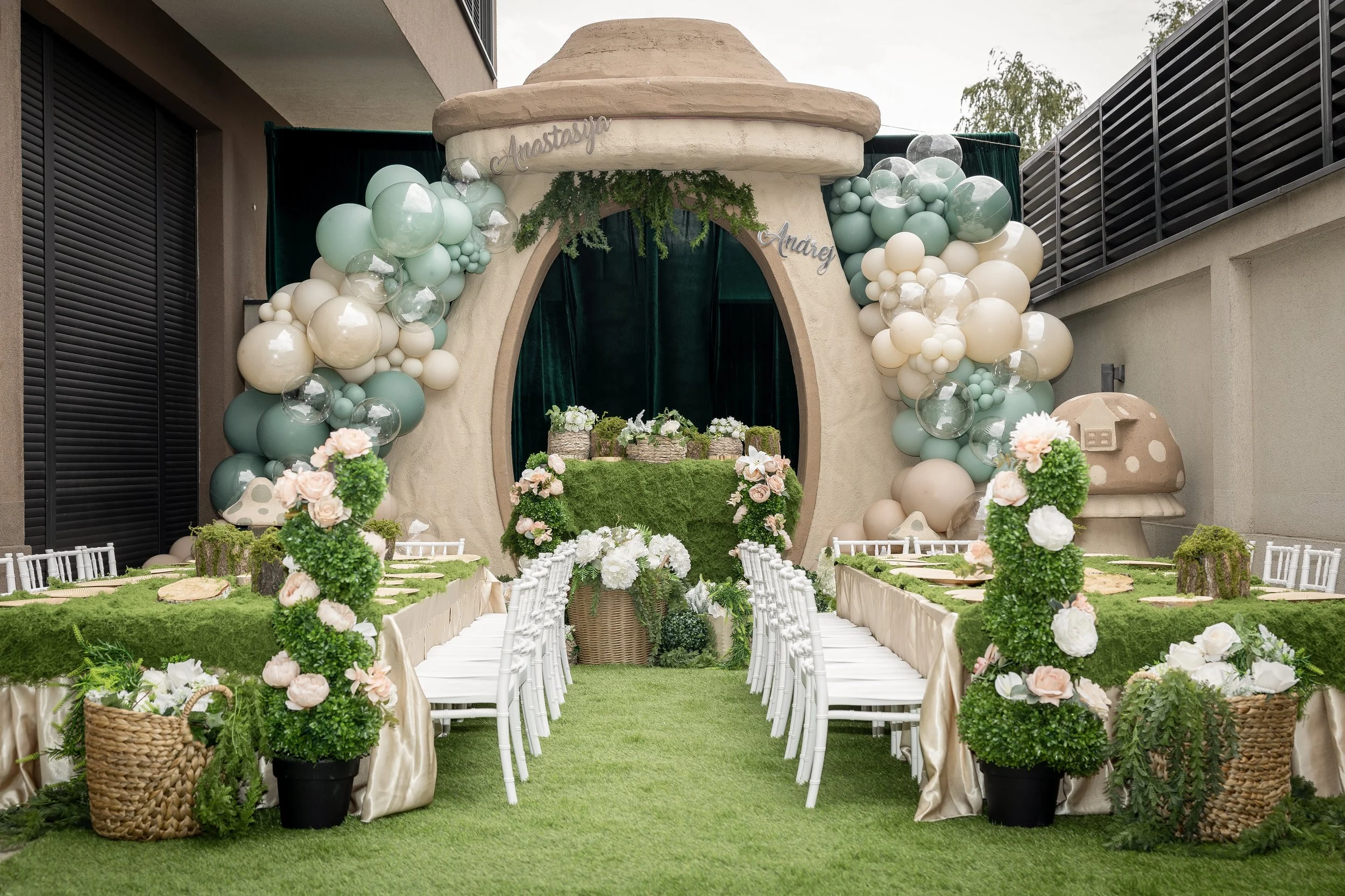 A whimsical, fairy-tale-themed outdoor wedding setup with a large rounded archway decorated with pastel green, beige, and clear balloons, green foliage, and fairy-tale inspired elements. There are long tables with beige tablecloths and white chairs, decorated with floral arrangements in baskets and potted greenery. The backdrop features a small, castle-like structure with a sign that reads 'Anastasia' and 'Andrey.'