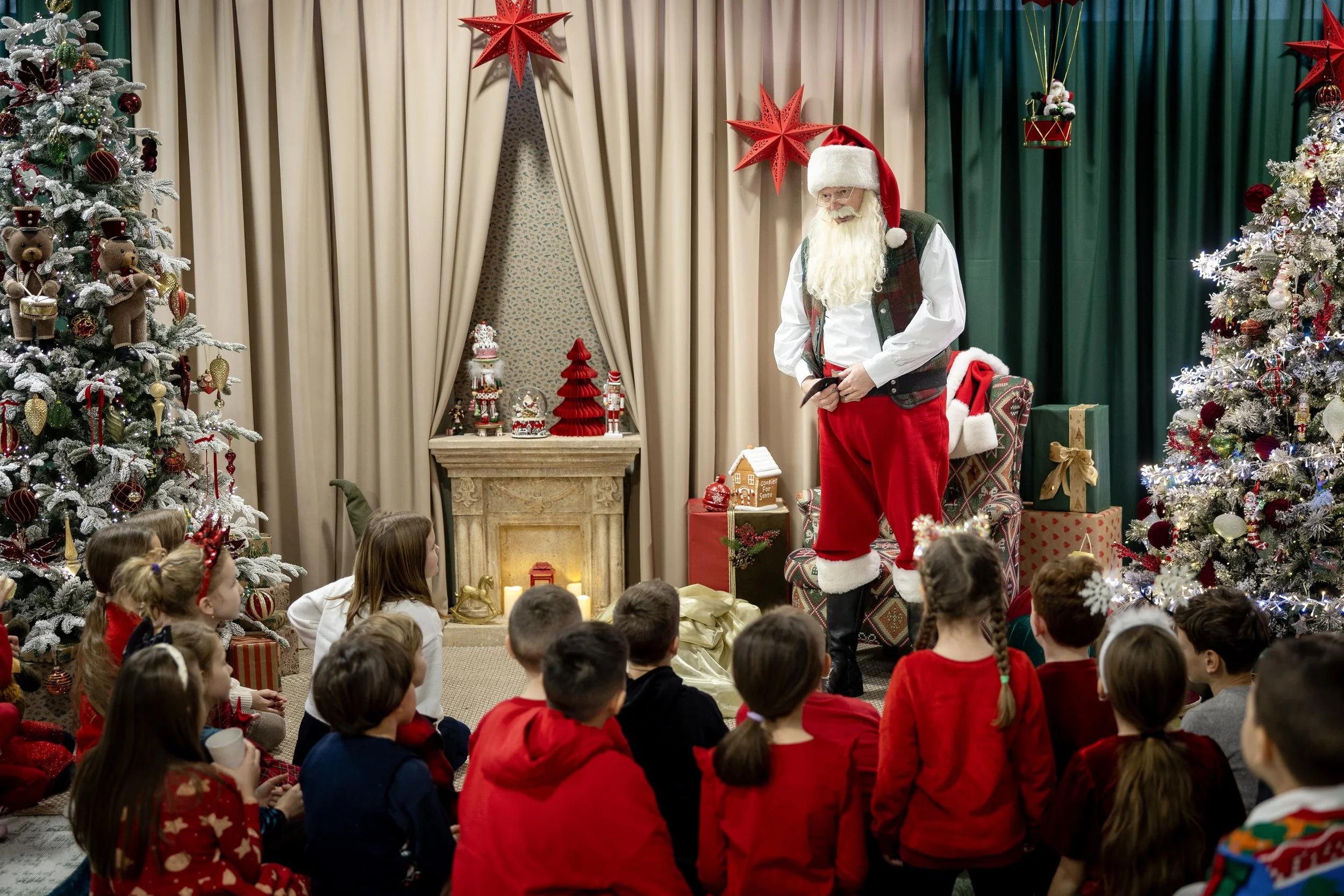 Santa Claus standing in front of children seated on the floor, surrounded by decorated Christmas trees and holiday decorations.