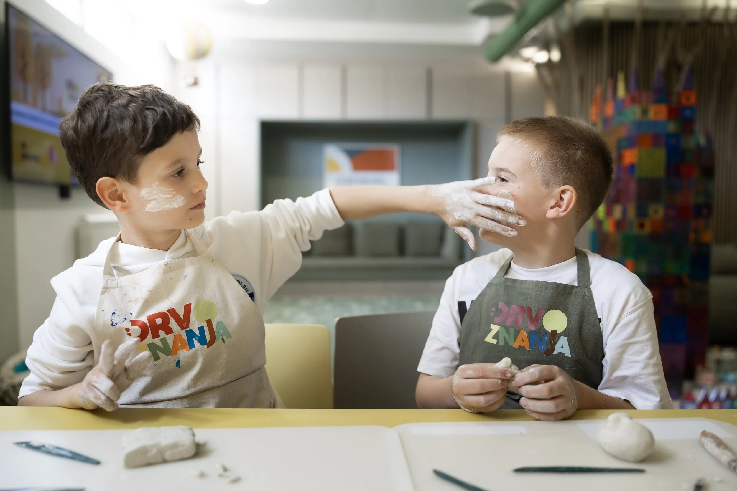 Two young boys with face paint and aprons, sitting at a table in a classroom, making clay sculptures with their hands.