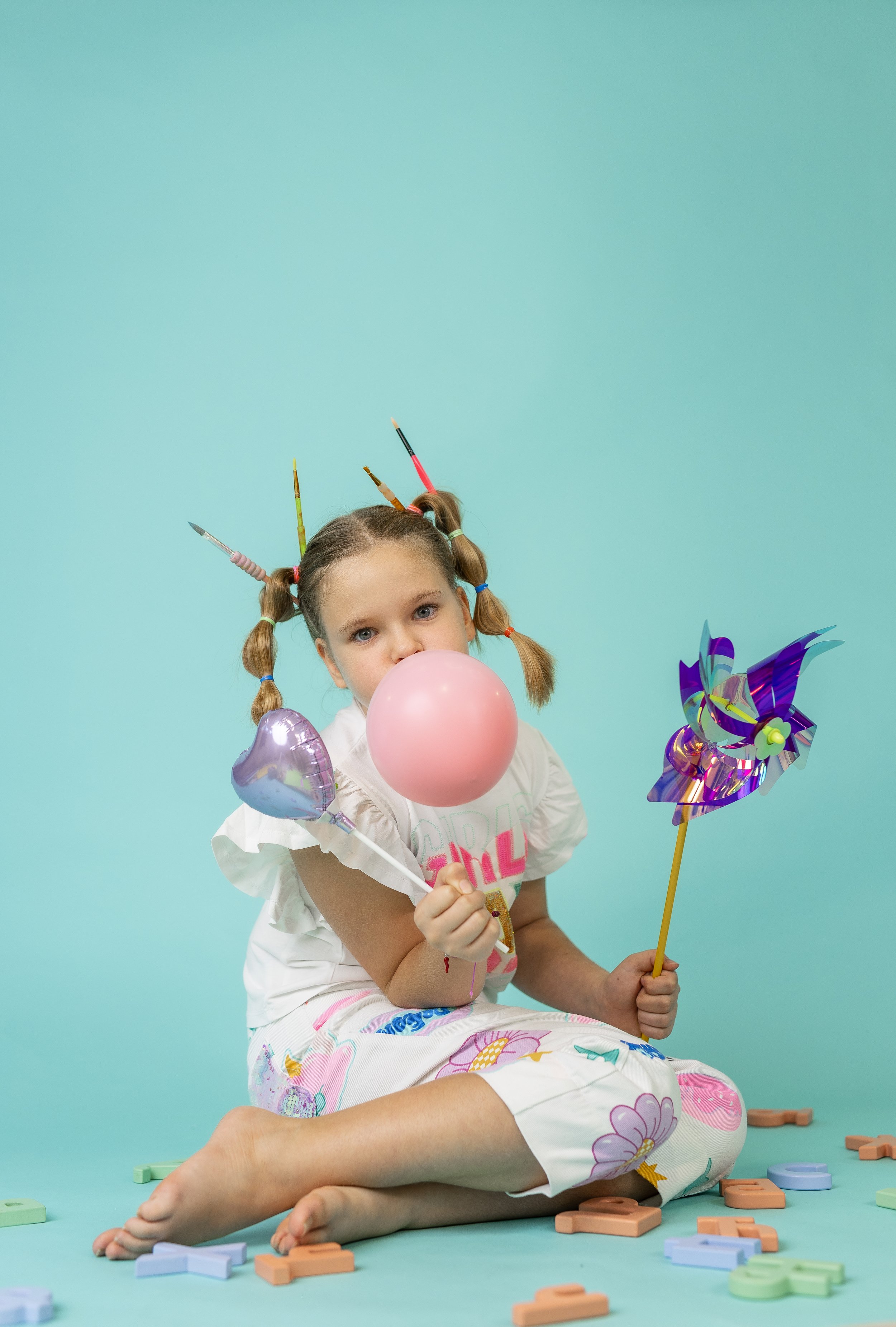A young girl with pigtails decorated with colored pencils plays with balloons, pinwheels, and puzzle pieces on a pastel teal background.