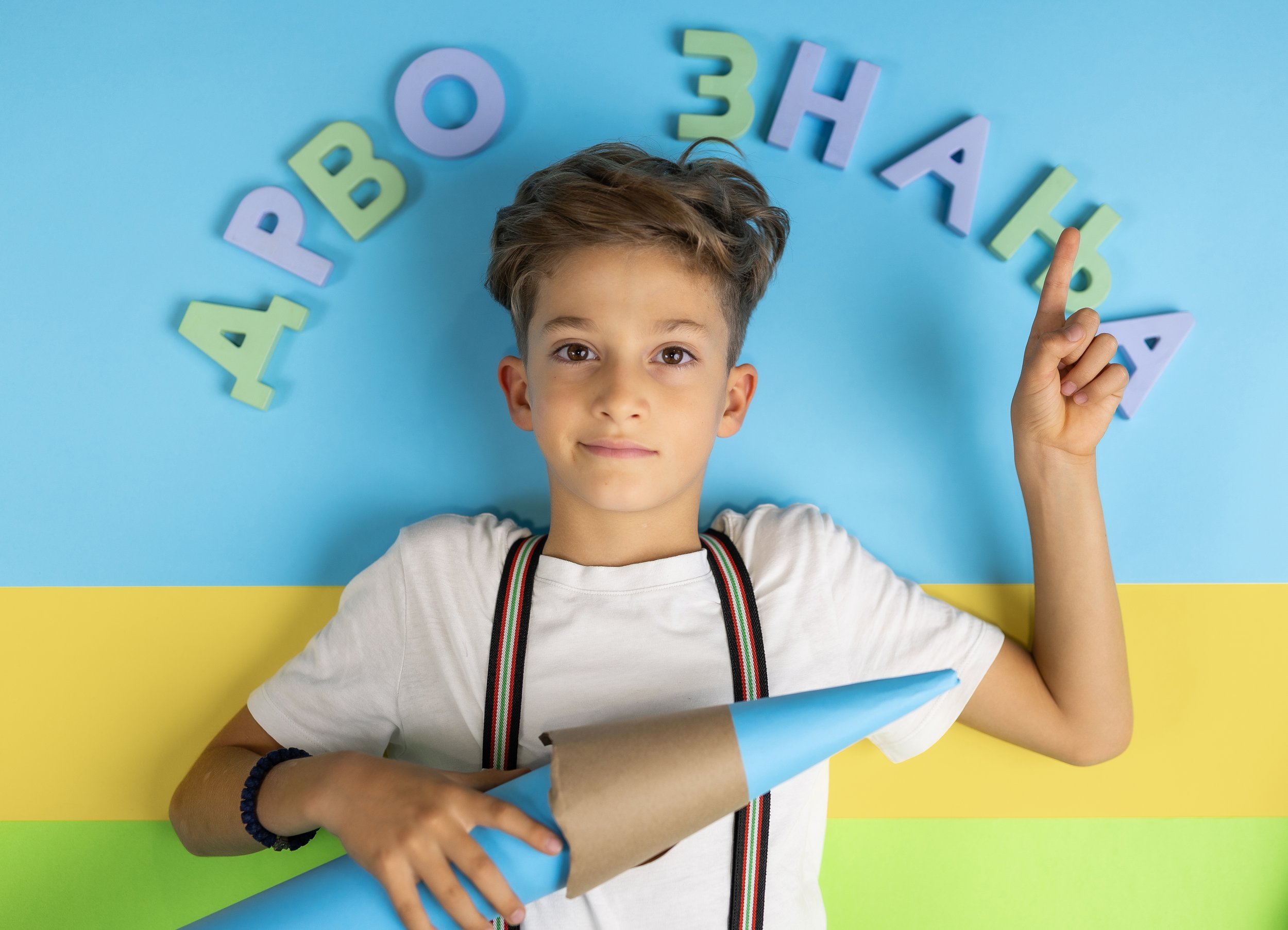A young boy with brown hair and wearing a white t-shirt, a colorful striped suspenders, and a black bracelet on his left wrist, lying on a background with horizontal blue, green, and yellow stripes, surrounded by colorful foam letters spelling 'HAPPY BIRTHDAY' arranged in an arc over his head, with his right index finger pointing upward.