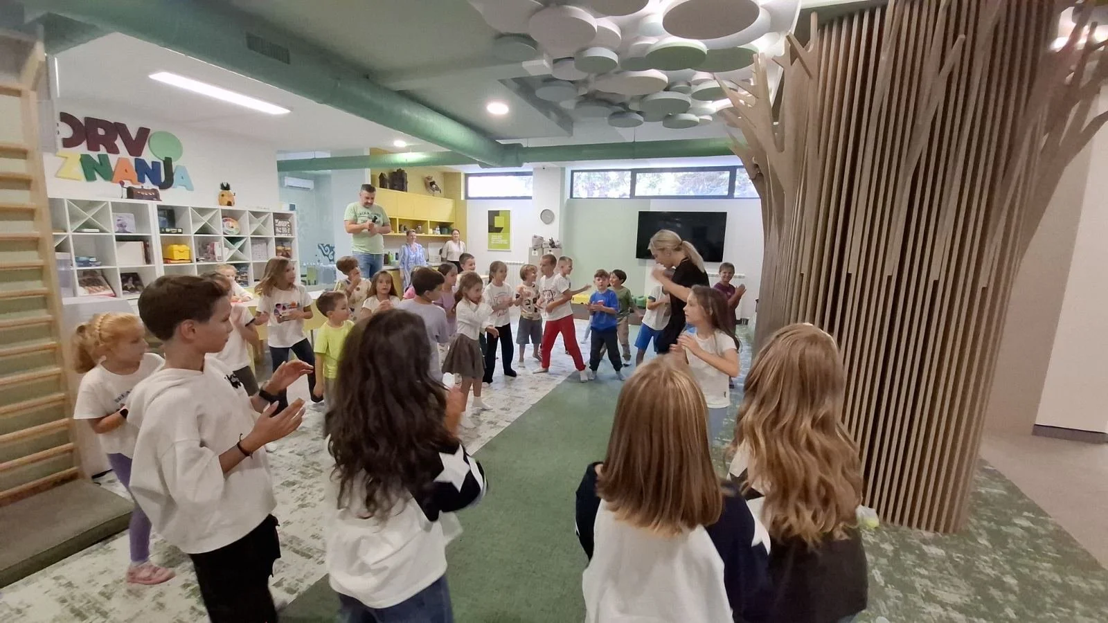 Children participating in an activity inside a room with shelves, a large tree sculpture, and a TV. A woman and a man are also present, and the room has playful, colorful decor.