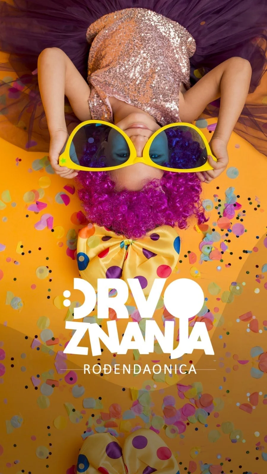 Child lying on yellow surface surrounded by colorful confetti, wearing a pink sequined dress, pink wig, large yellow sunglasses, and a yellow polka-dotted bowtie.