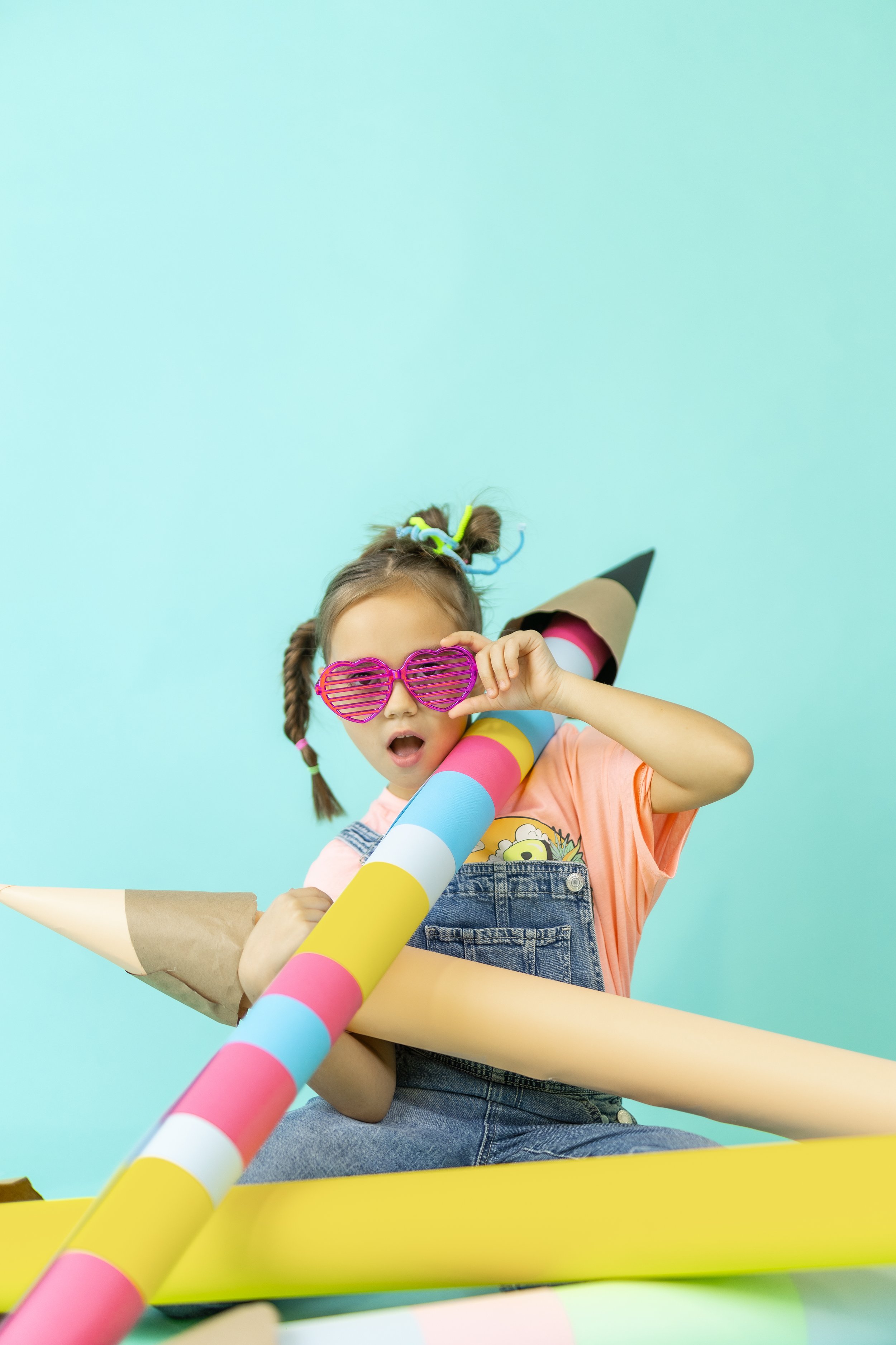 A young girl with braided hair, wearing heart-shaped pink sunglasses and a pink shirt with graphic on it, sitting on a yellow surface against a blue background, surrounded by colorful striped paper and cardboard cones.