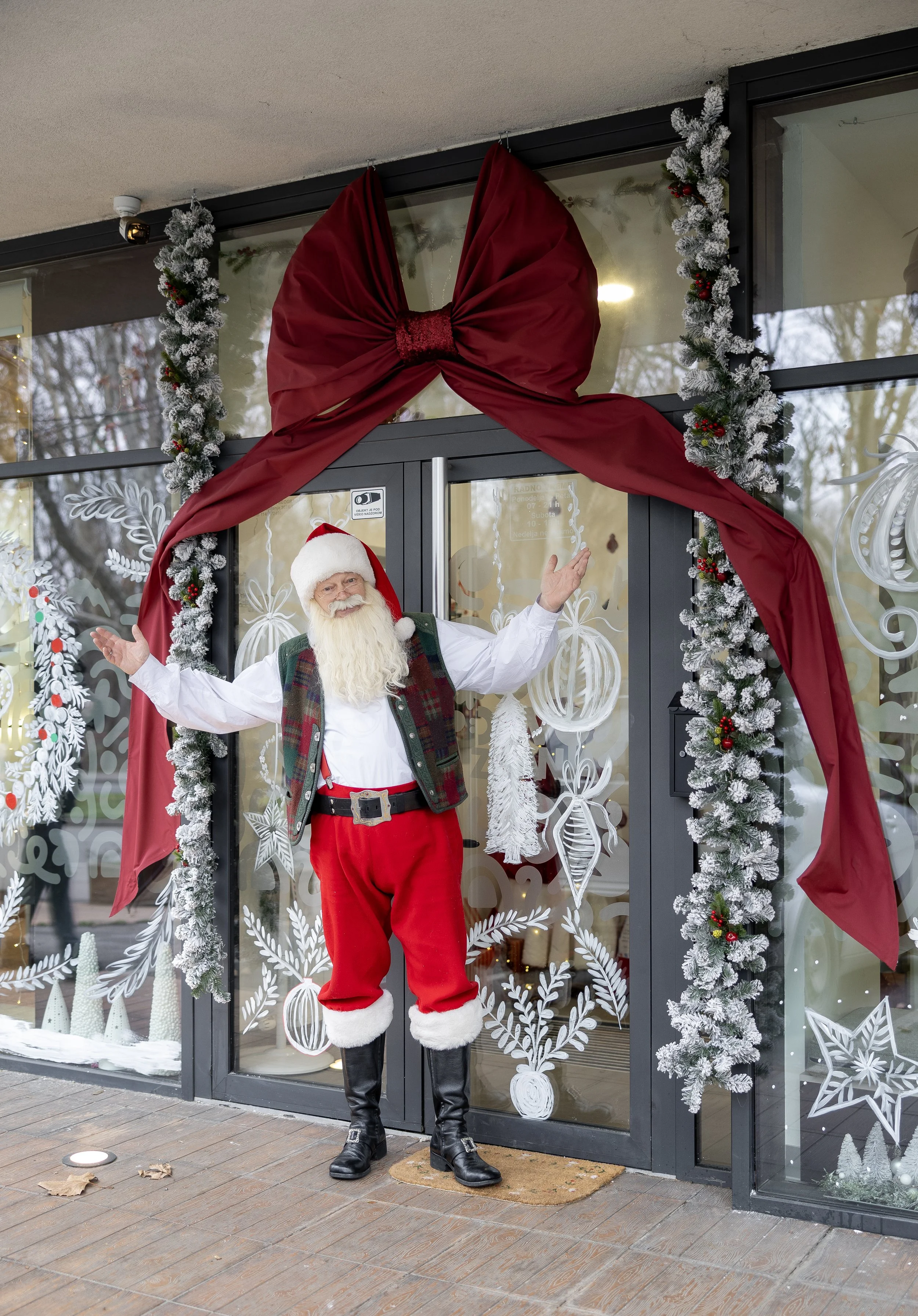 Santa Claus standing outdoors in front of a festive winter display, wearing a red hat with white fur trim, a white shirt, red pants, black boots, and a green plaid vest. He is smiling and waving with both hands.